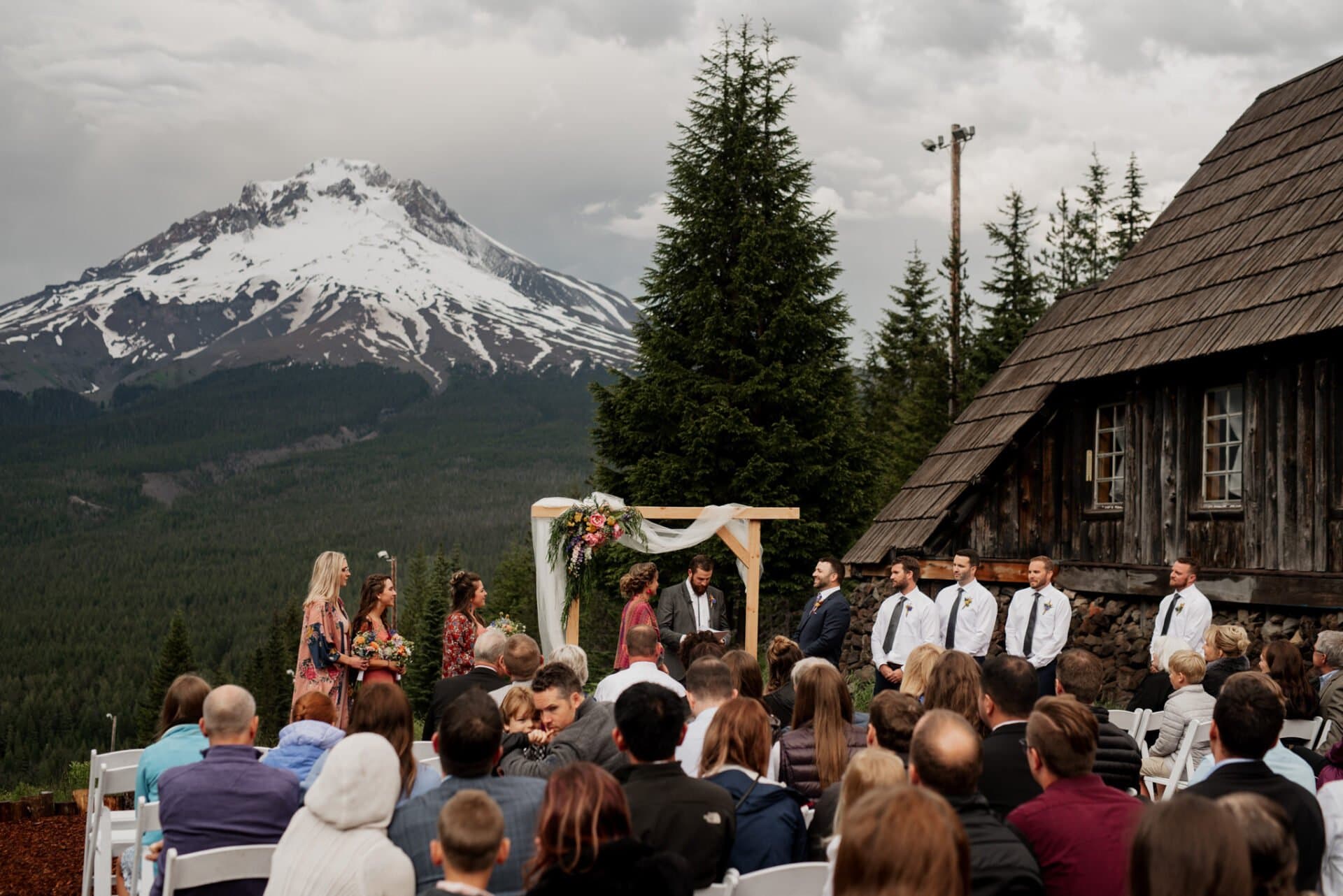 A wedding ceremony unfolds in front of a wooden cabin with snow-capped mountains framing the scene, reminiscent of a Skibowl backdrop. The bride and groom stand under a flower-adorned arch, surrounded by the wedding party and guests seated on white chairs.