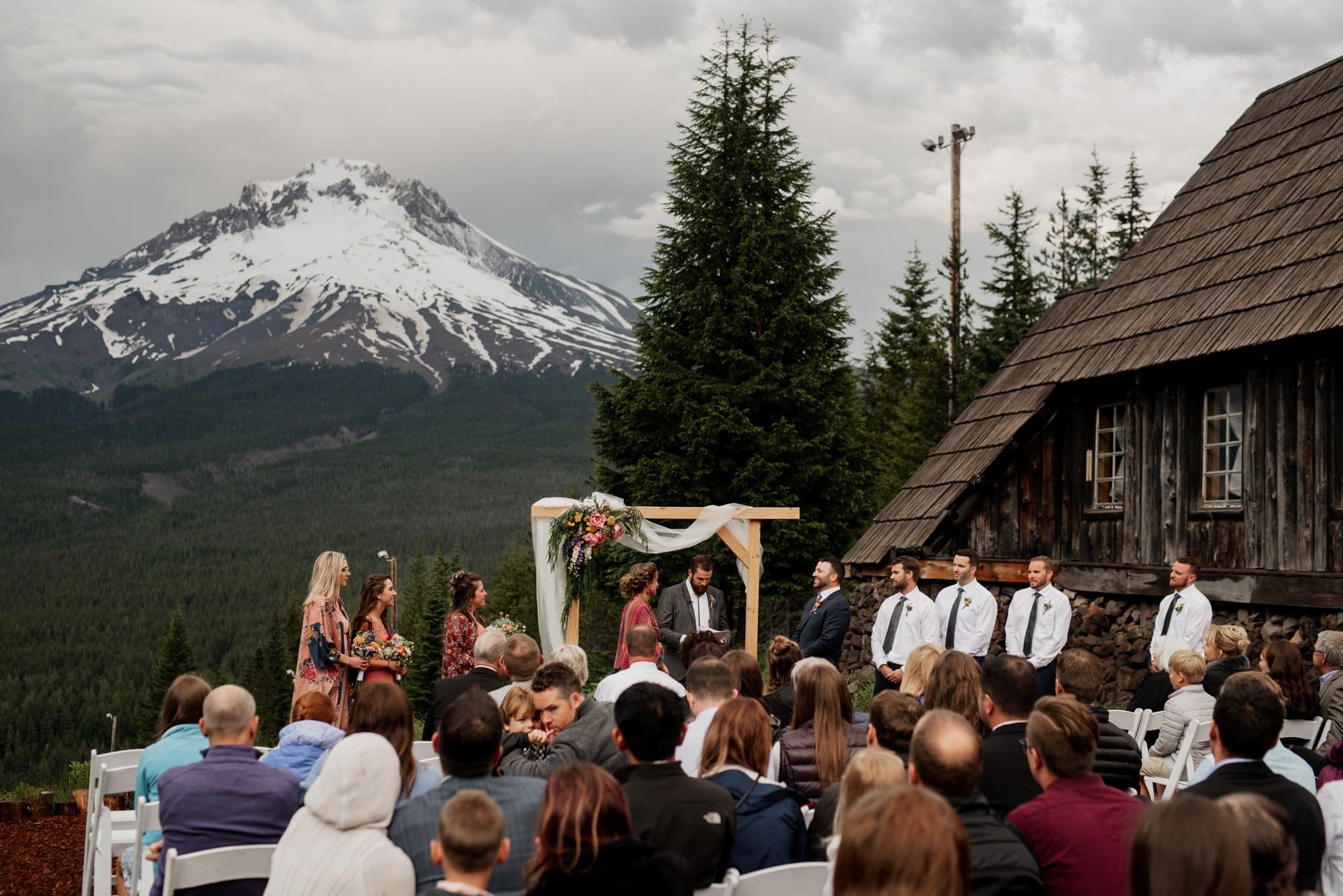 A wedding ceremony unfolds in front of a wooden cabin with snow-capped mountains framing the scene, reminiscent of a Skibowl backdrop. The bride and groom stand under a flower-adorned arch, surrounded by the wedding party and guests seated on white chairs.