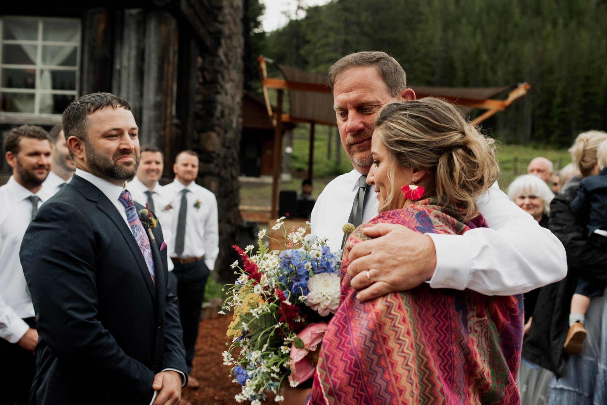 A bride wrapped in a colorful shawl holds a bouquet and is embraced by her father, both surrounded by formally dressed onlookers outdoors. A suited groom watches as the rustic charm of Mt Hood Ski Bowl provides a picturesque backdrop to their heartfelt wedding moment.