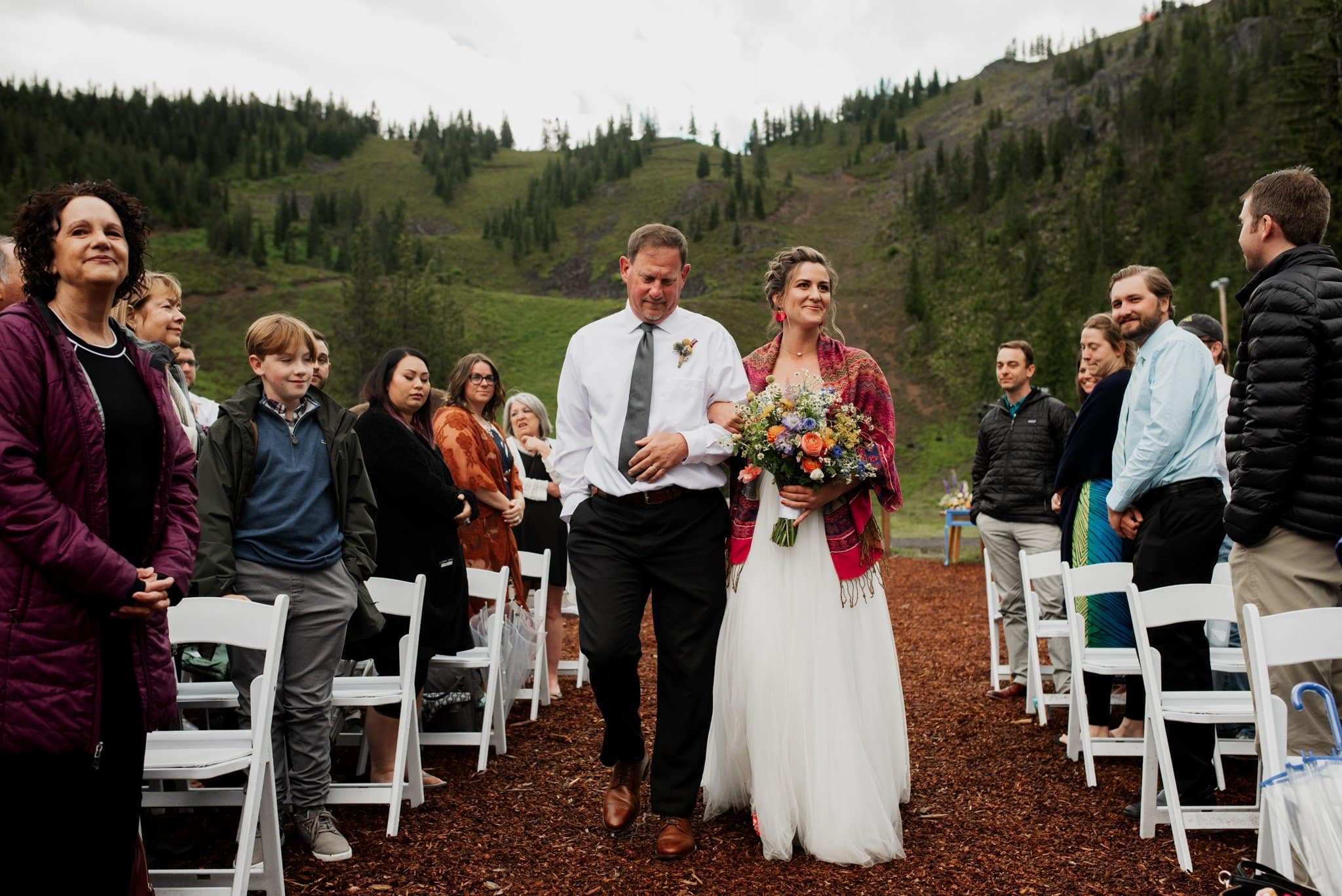 A bride in a white dress and colorful shawl walks down an outdoor aisle at Mt Hood Ski Bowl with a man in a white shirt and tie. Guests smile while standing on either side. The background features green hills and a cloudy sky, adding to the scenic wedding venue charm.