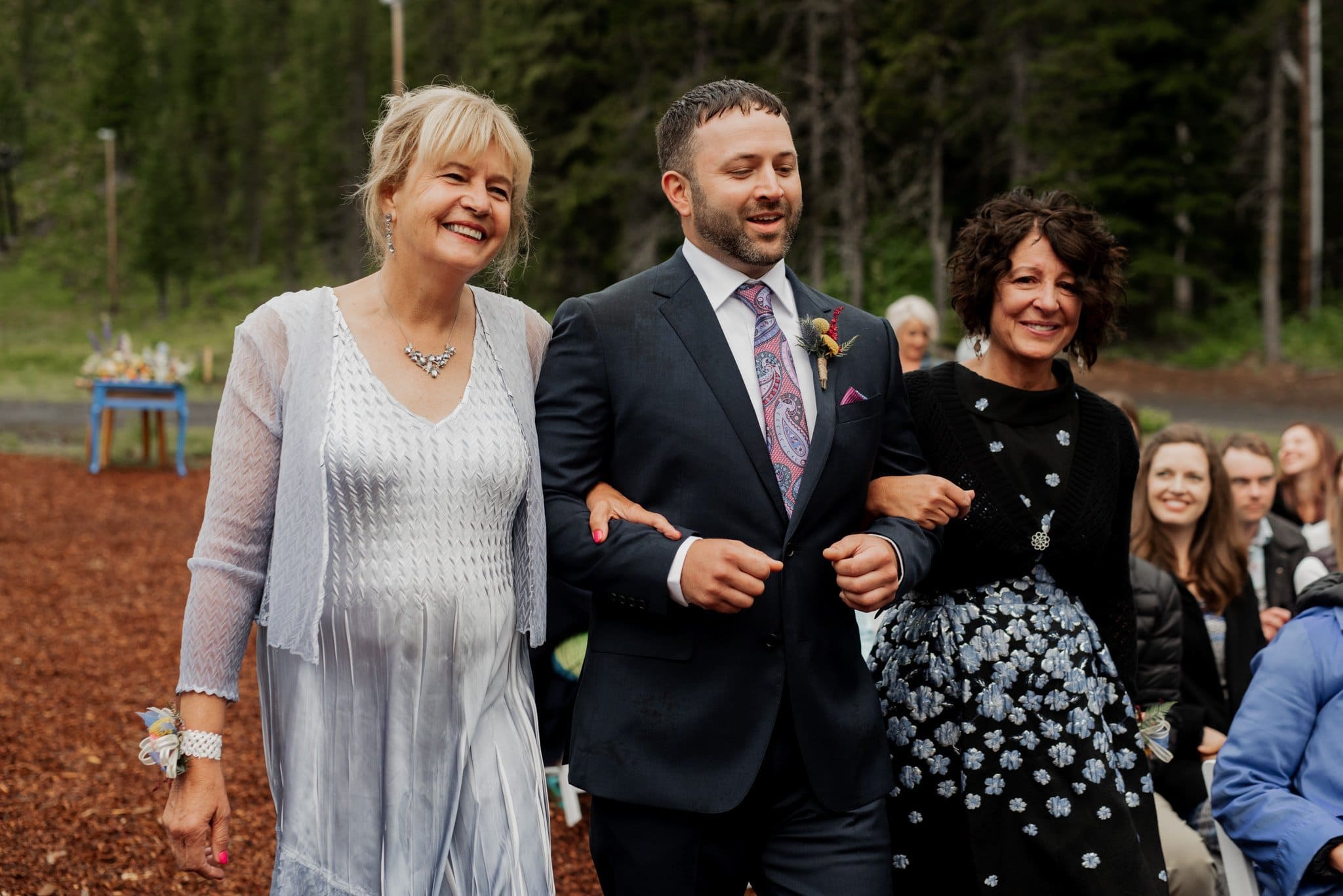 A smiling groom in a dark suit walks arm-in-arm with two women in dresses down an outdoor aisle covered in wood chips at the picturesque Mt Hood Ski Bowl. Surrounded by trees, guests are seated in the background, watching the charming procession unfold.