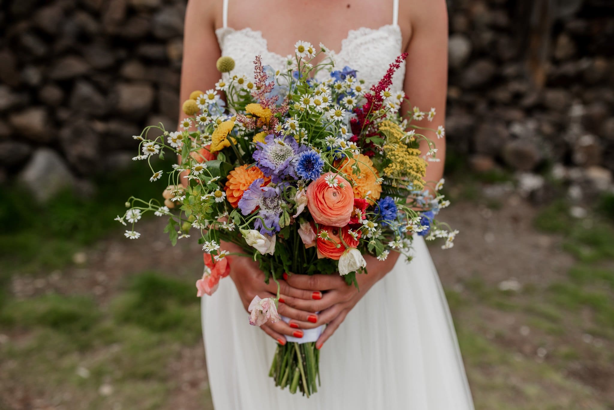 A bride in a white lace dress holds a vibrant bouquet of mixed flowers, featuring orange, purple, blue, yellow, and white blooms. Against the rustic stone wall and green grass at Mt Hood Ski Bowls breathtaking venue, her nails are painted orange.