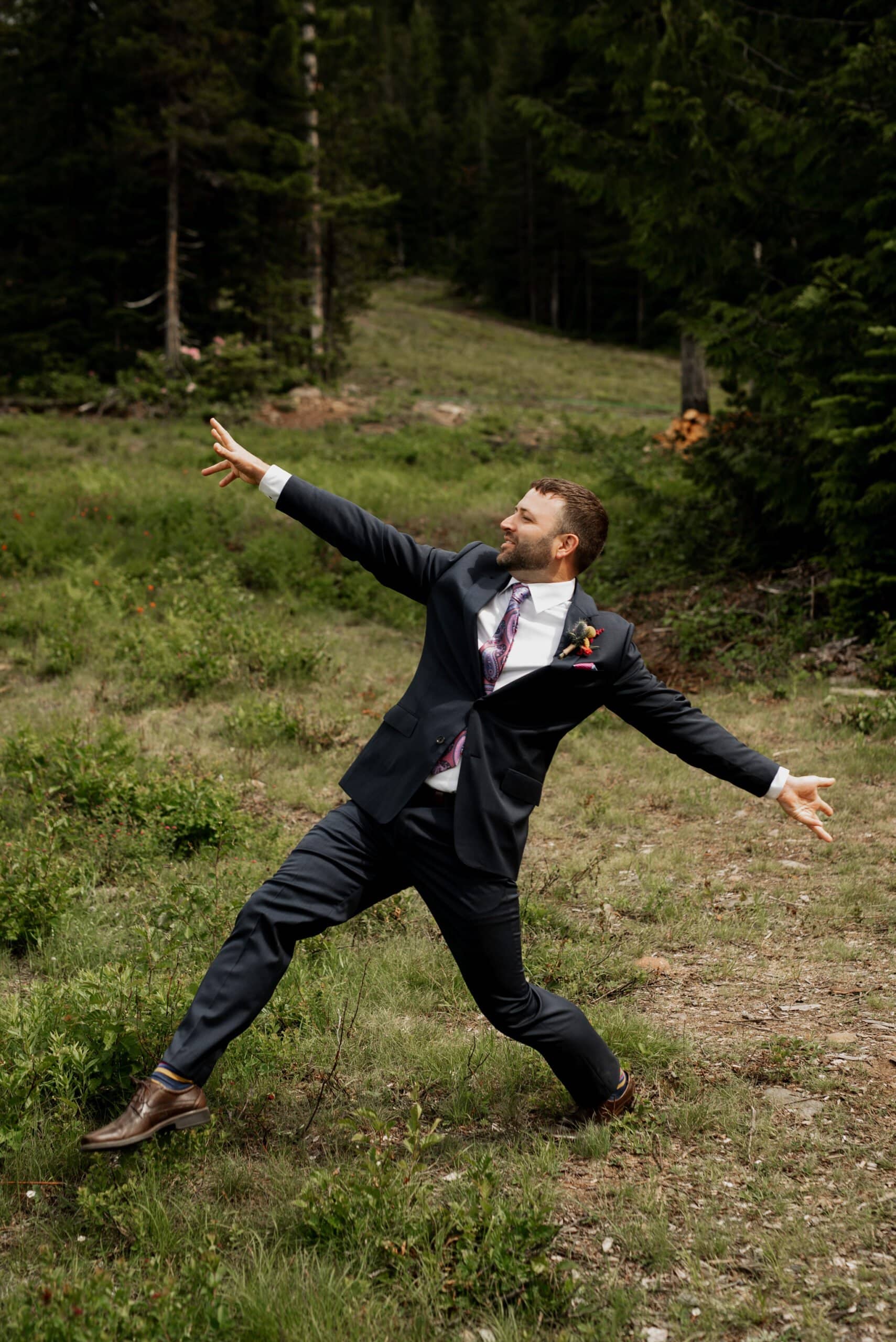 A groom in a suit strikes a dramatic pose amid the lush greenery of a grassy, wooded area at Mt Hood Ski Bowl. With one arm reaching skyward and the other out to the side, he leans back, creating a dynamic appearance—a scene worthy of a wedding moment.