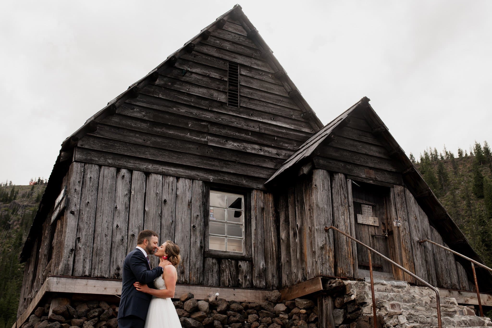 A couple in wedding attire lovingly embrace in front of an old, rustic wooden warming hut with a peaked roof, nestled near Mt Hood Ski Bowl. The overcast sky and dense forest of evergreens create a serene backdrop for their special day.