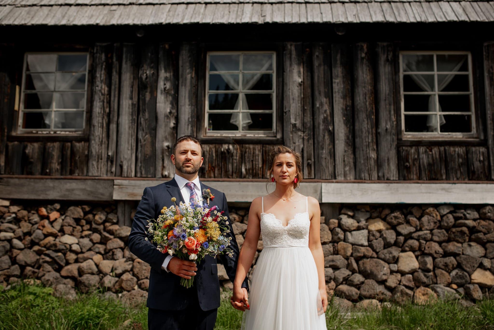 Newlyweds stand holding hands in front of a rustic wooden warming hut with a stone foundation at Mt Hood Ski Bowl. The man, in a suit, holds a colorful bouquet while the woman wears a white wedding dress. Their serious expressions blend with the backdrop of windows and lush grass.