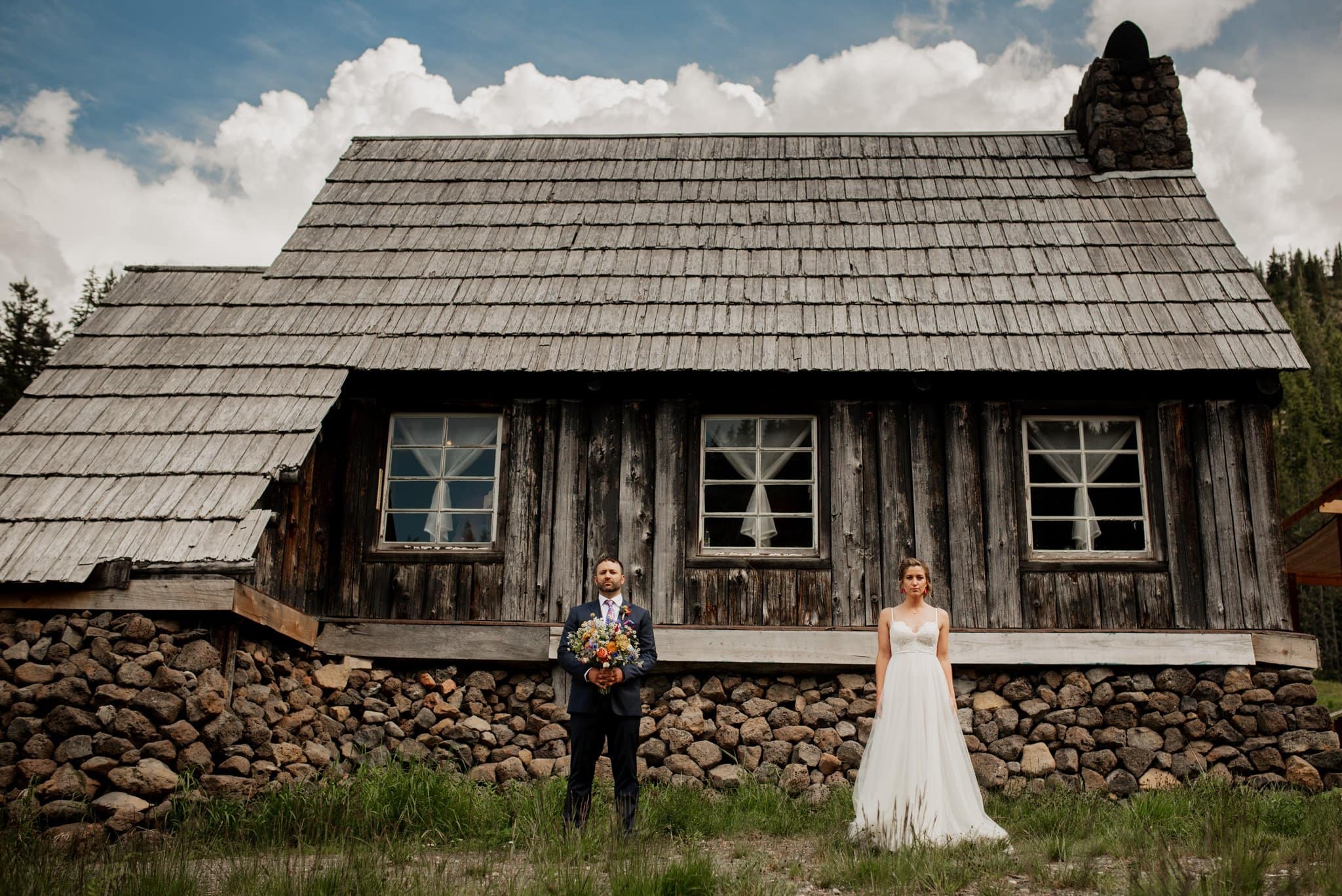 A bride and groom stand side by side in front of the rustic Mt Hood Skibowl warming hut, with a stone foundation. The groom holds a bouquet, and the bride wears a flowing white dress. A cloudy sky and green trees complete this picturesque scene.