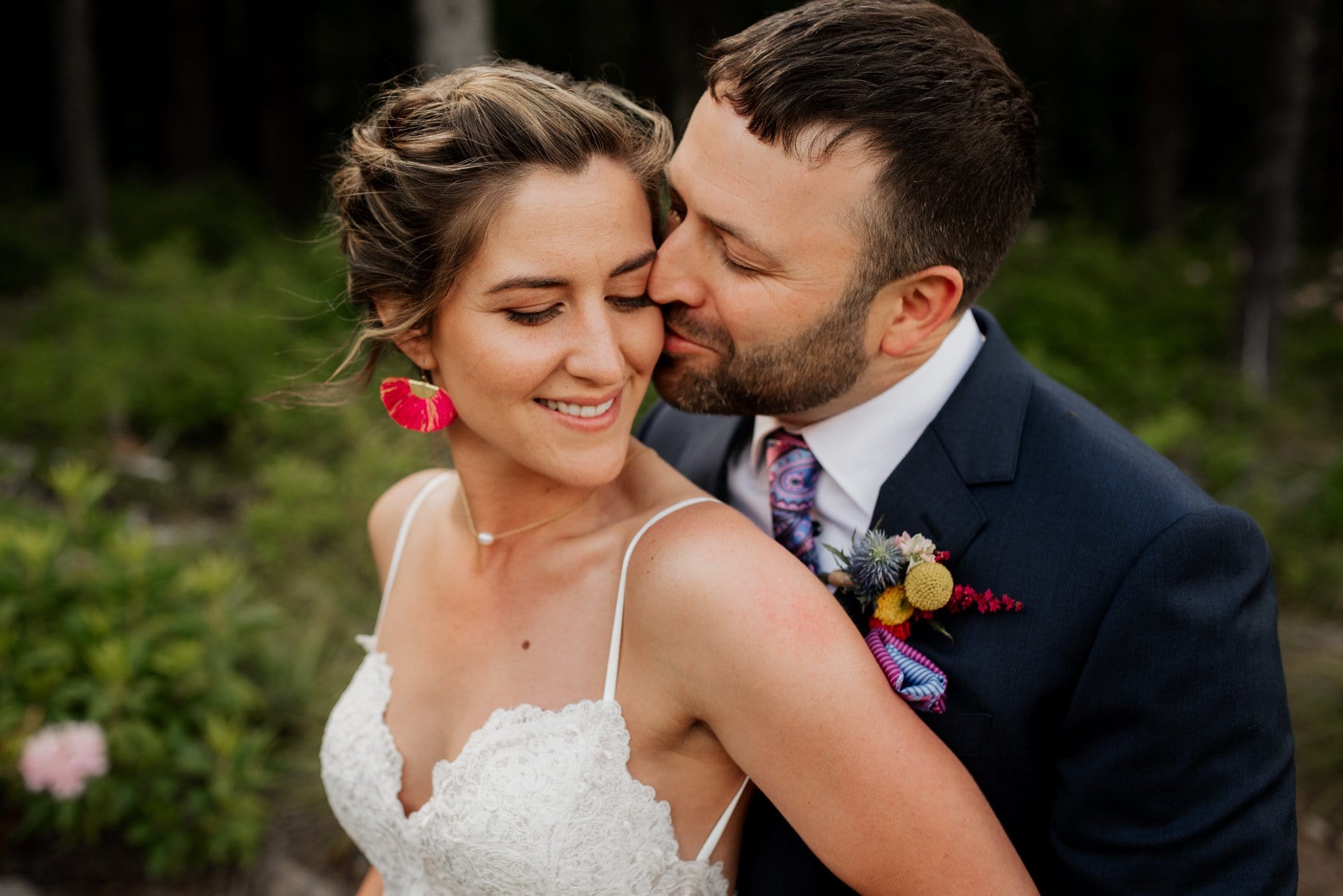 A groom in a navy suit lovingly kisses a smiling bride on the cheek during their Mt. Hood Ski Bowl wedding. The bride, in a white lace dress and pink earrings, holds a bouquet. They stand outdoors with greenery surrounding them, capturing a joyful and intimate moment.
