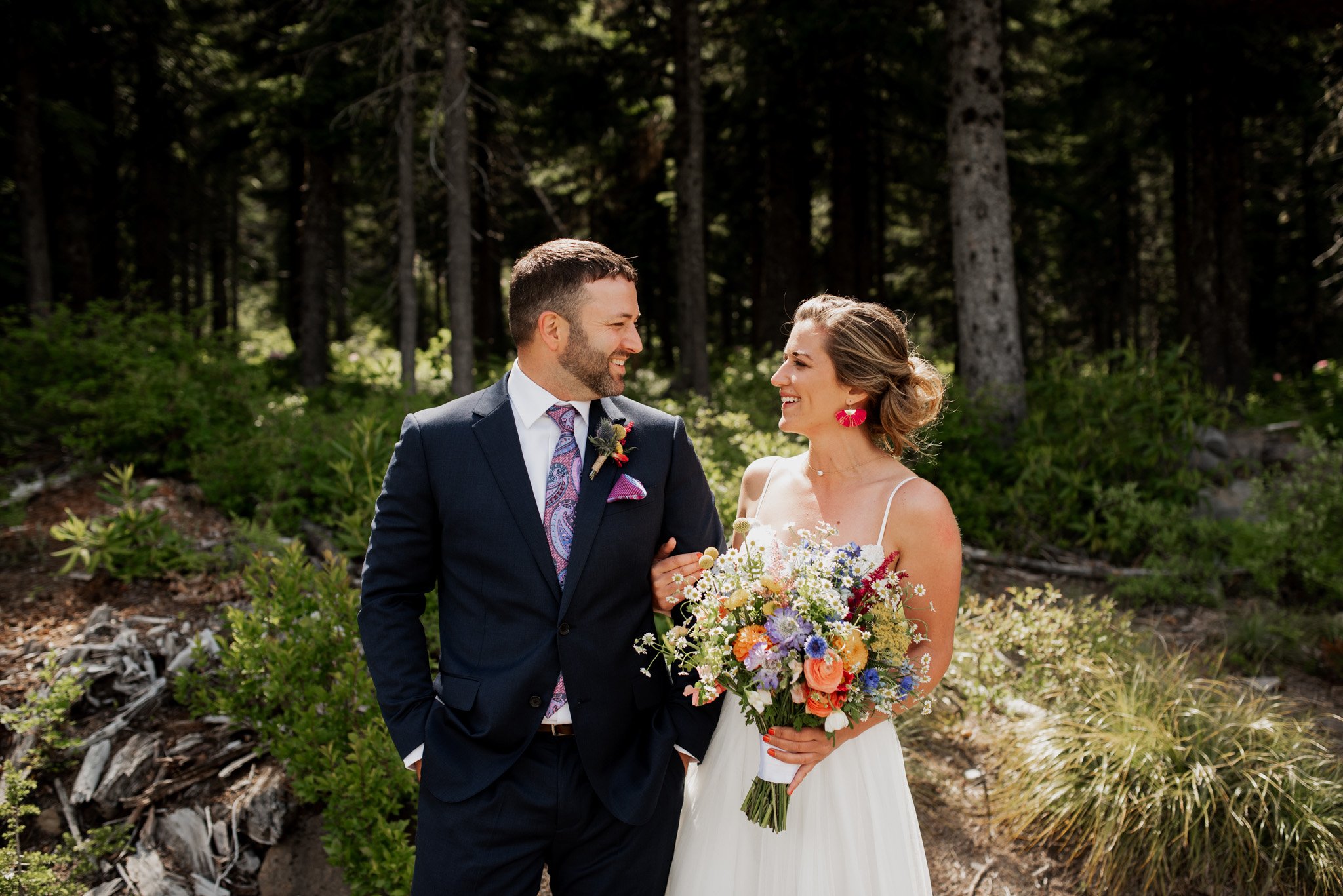 A bride and groom stand arm in arm in a forest at Mt. Hood Ski Bowl. The bride holds a colorful bouquet and wears bright earrings with her white dress; the groom sports a navy suit with a vibrant tie. Smiling at each other, they are enveloped by lush greenery, capturing their perfect moment.