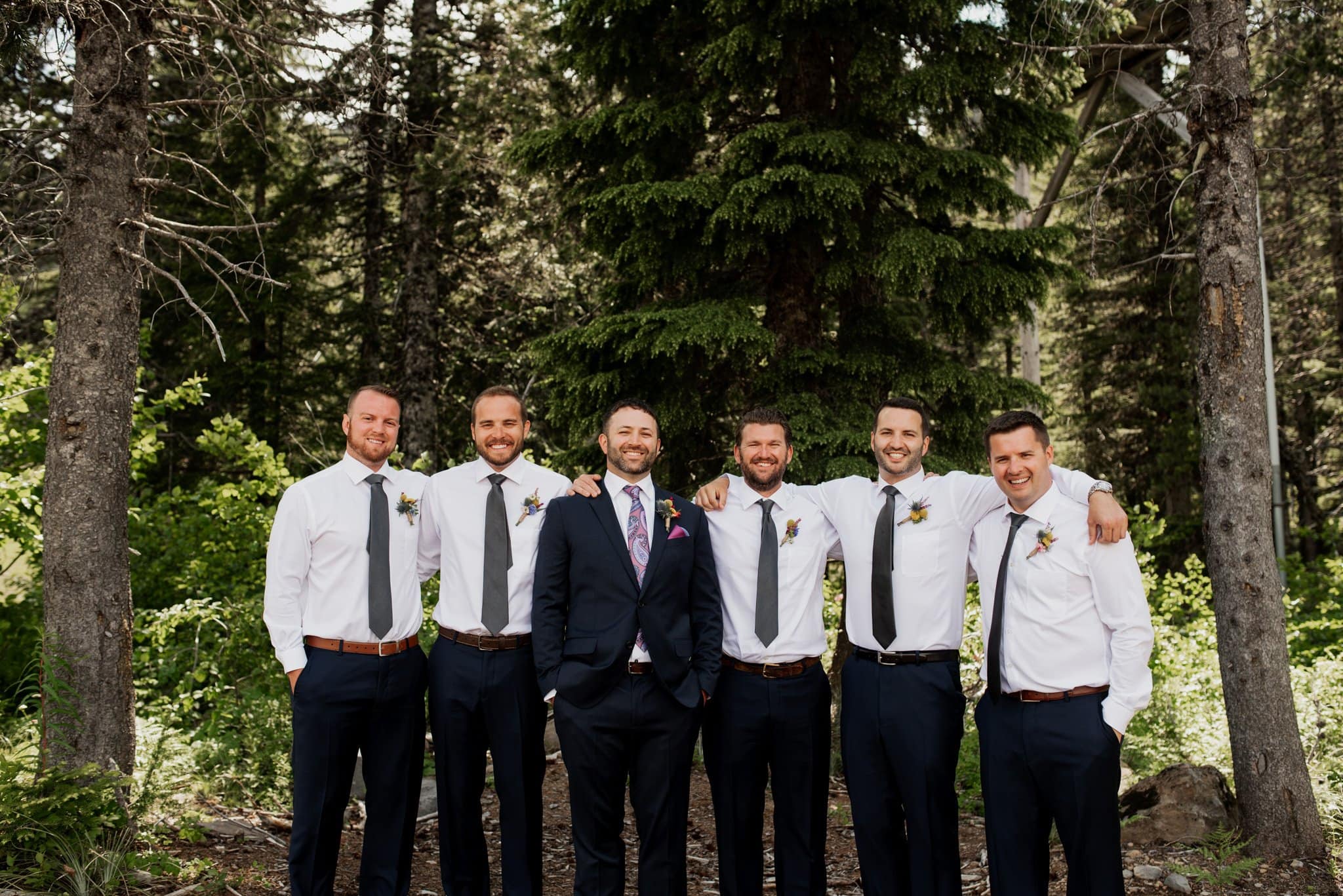 Six men stand in a forest setting with trees behind them, at a Mt Hood Ski Bowl wedding. Five wear white shirts and dark pants, while the one in the center sports a dark suit and patterned tie. All are smiling, capturing the essence of this joyous formal event.