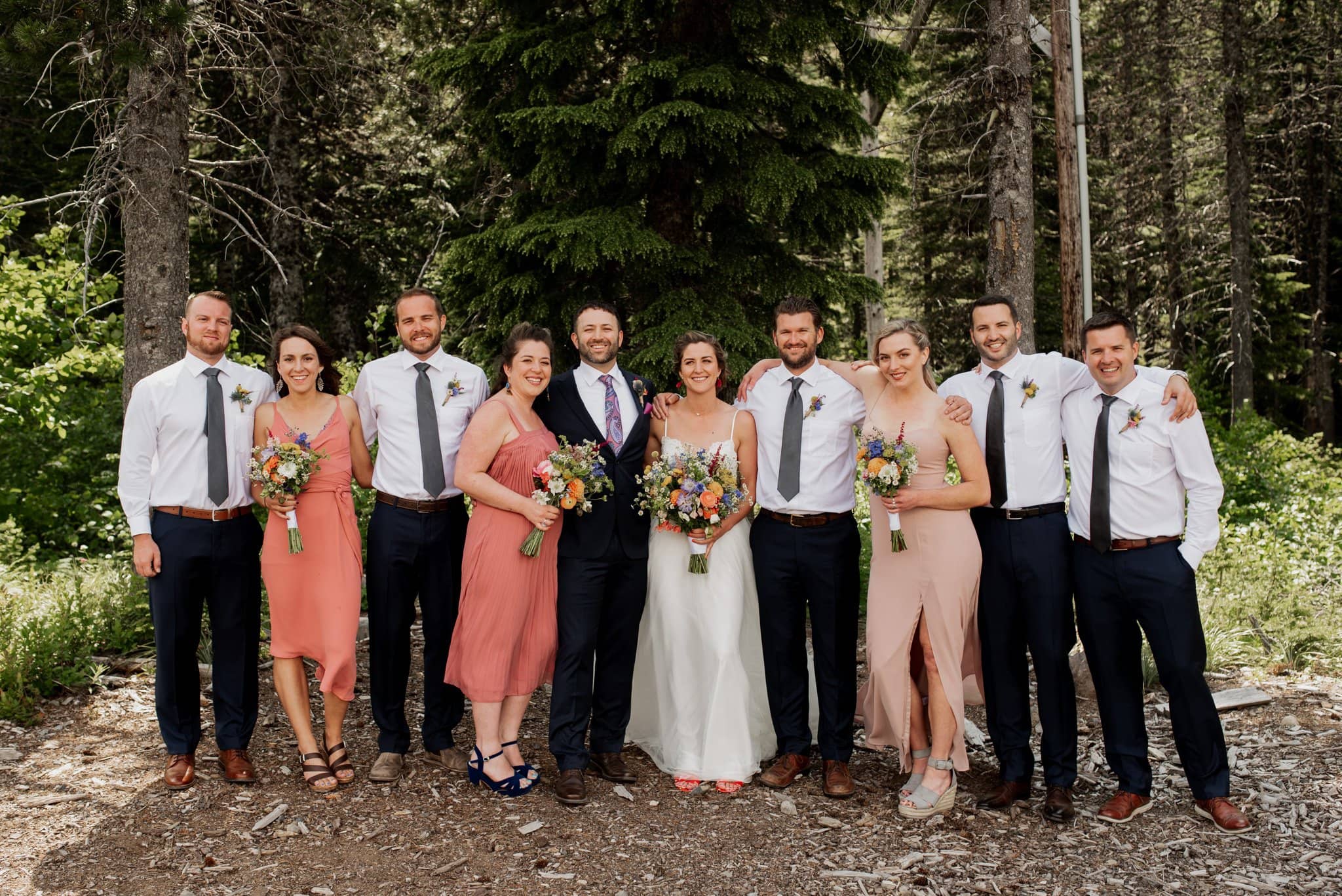 A wedding party poses in the enchanting forest of Mt Hood Ski Bowl. The bride, in a white dress with a colorful bouquet, stands beside the groom in a dark suit. Bridesmaids wearing coral and blush dresses and groomsmen in white shirts with dark pants surround the couple beautifully.