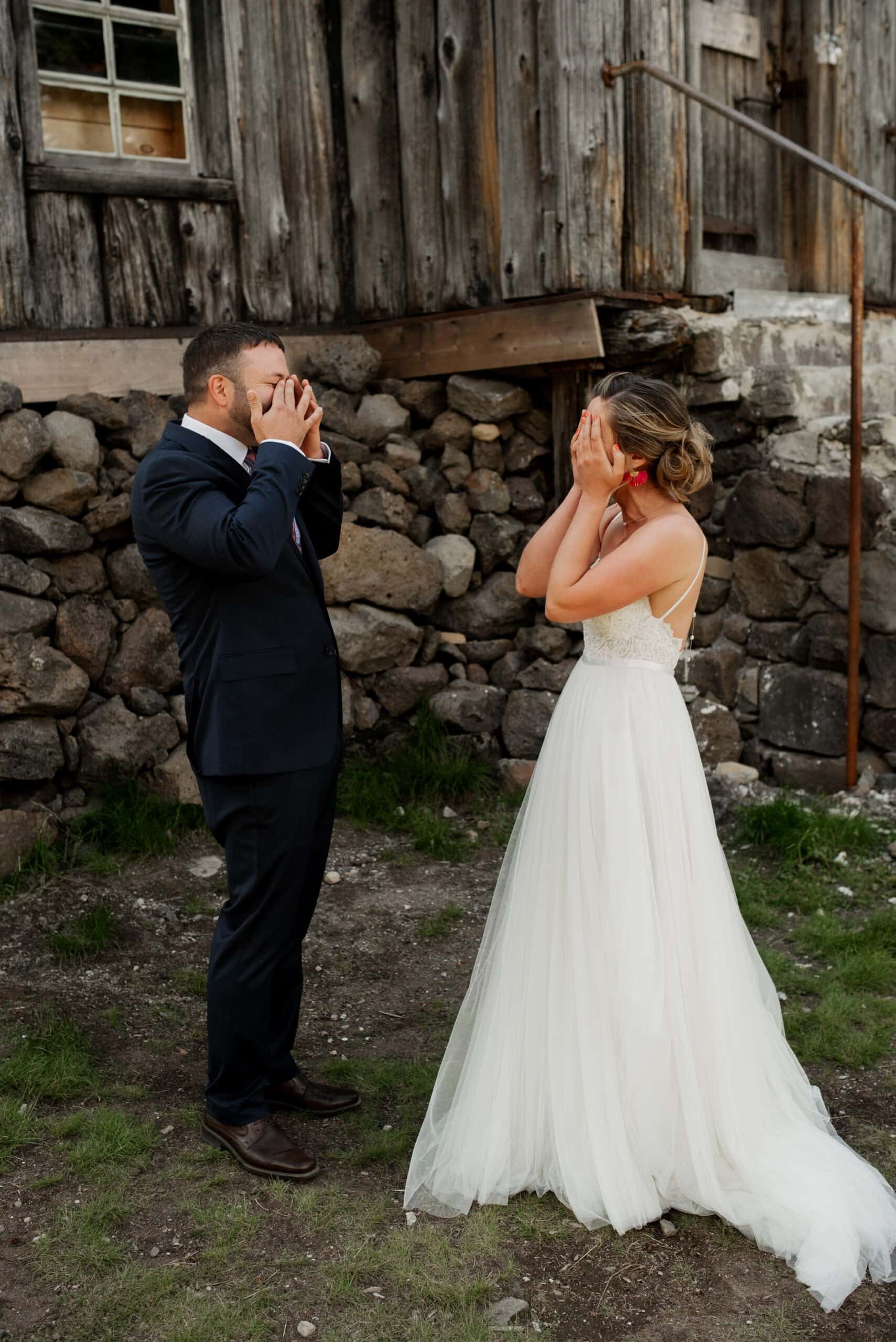 A bride in a white dress and a man in a blue suit stand outdoors at Mt Hood Ski Bowl, covering their faces with their hands. They appear surprised or emotional, set against the backdrop of a rustic stone and wooden warming hut.