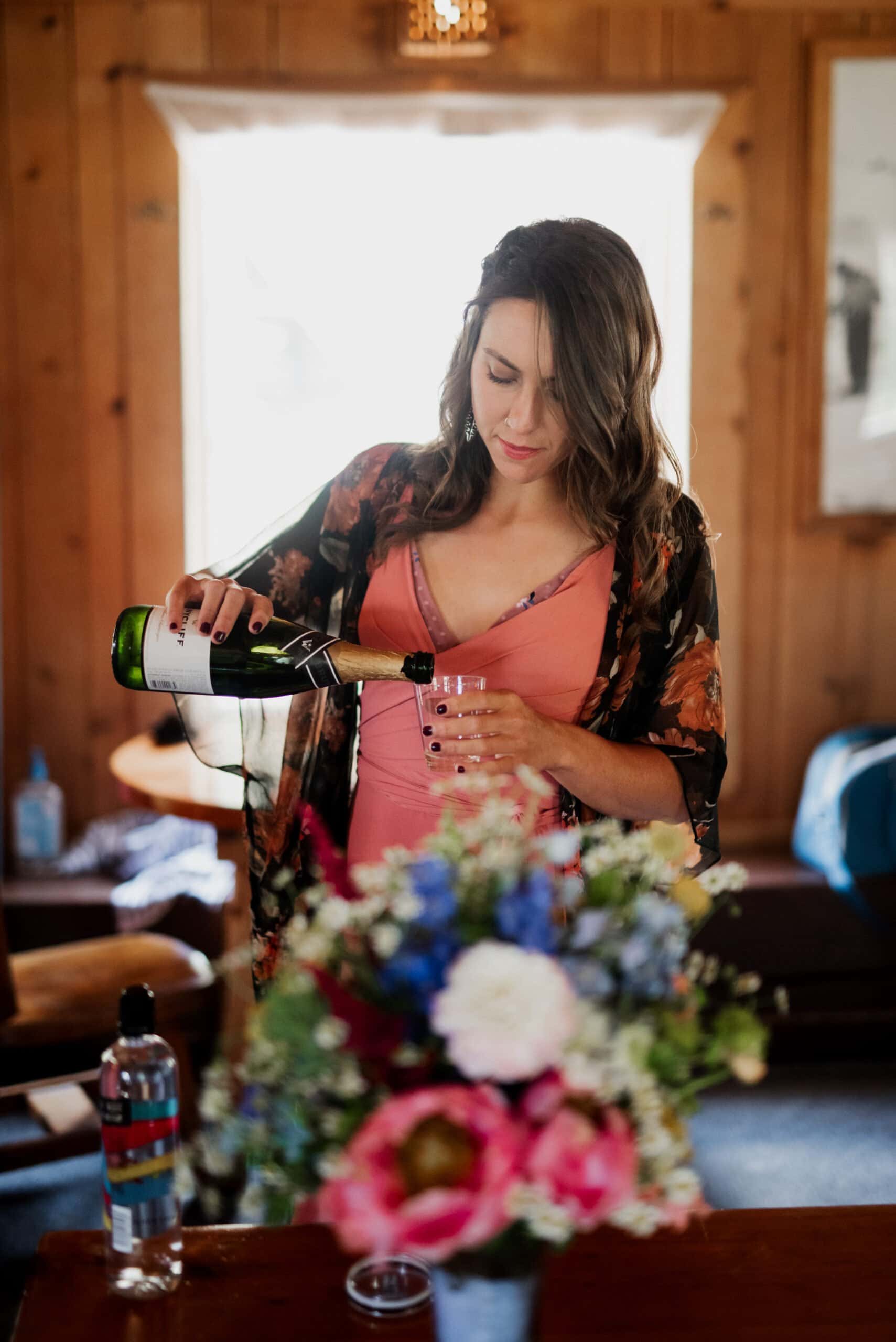 In a cozy room at Mt Hood Skibowl, a woman in a floral robe pours champagne into a glass. She stands by a wooden table adorned with a colorful bouquet and water bottle, against wood-paneled walls and an invitingly open window, setting the perfect scene for an intimate wedding moment.