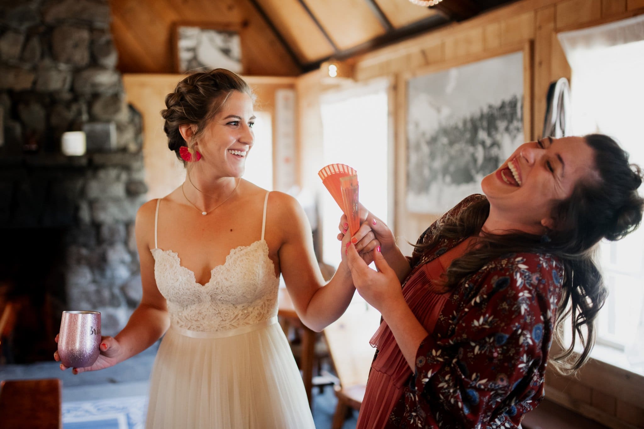Two women stand in a cozy room with wooden walls. One wears a white dress, holding a pink cup, while the other, in a colorful jacket, laughs heartily. They are getting ready for a Mt Hood Ski Bowl wedding, radiating joy beside the stone fireplace.