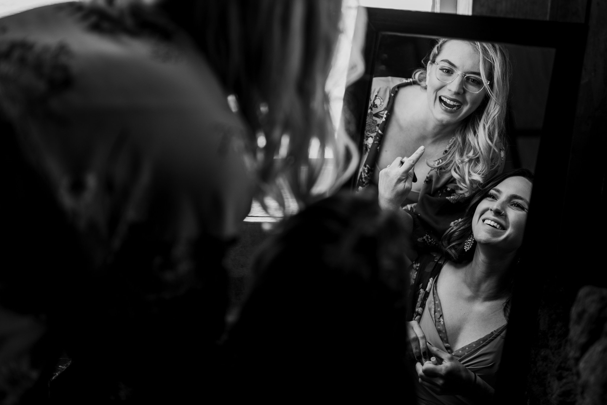 A black and white image captures two women laughing together as they prepare for a Mt Hood Ski Bowl wedding. One woman is applying lipstick, while the other adjusts her glasses, both bathed in light streaming from behind, illuminating their joyful expressions.