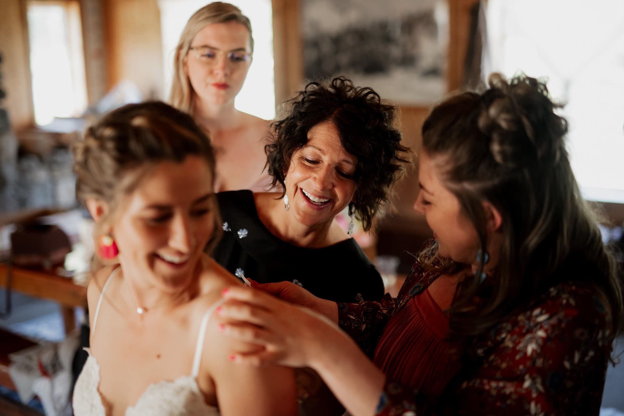 In a warmly lit room at Mt Hood Ski Bowl, four women are joyfully helping each other get ready. One in a white dress is assisted with her strap by another, while the remaining two smile and look on. The atmosphere is lighthearted and friendly.
