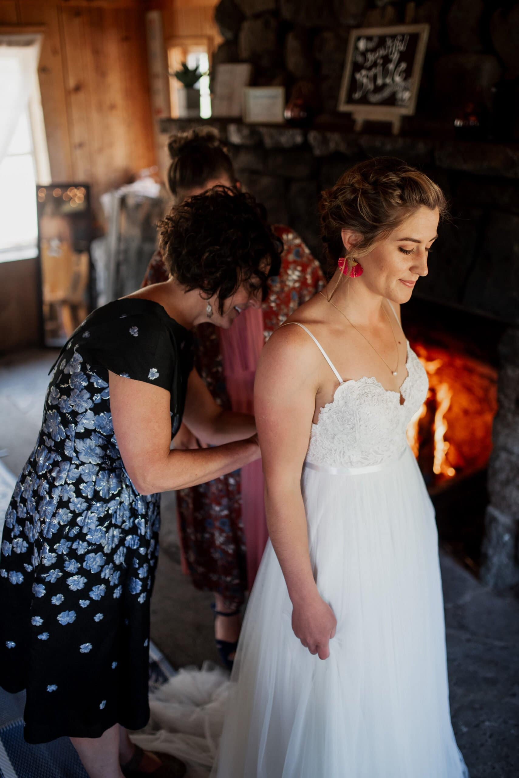 In a cozy, rustic setting reminiscent of a Mt Hood Ski Bowl wedding, a bride in a white dress stands smiling as two women adjust her gown. Indoors, with a roaring fire in the stone fireplace, she flaunts short hair and red earrings.