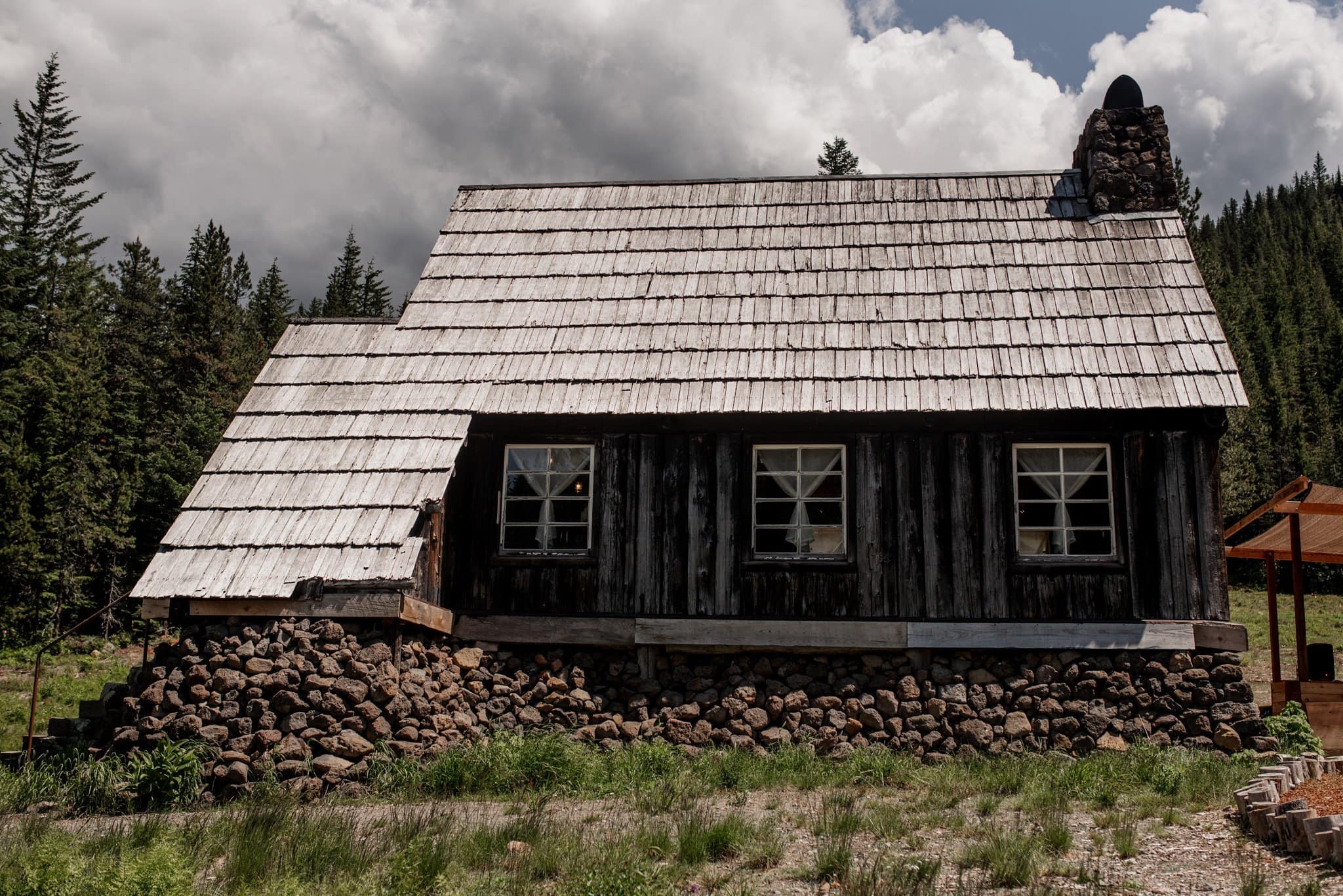 A rustic cabin with a weathered wooden exterior and stone foundation stands against a forested backdrop. Like a warming hut, its shingled roof and chimney exude coziness. A cloudy sky looms overhead, enhancing the remote, serene vibe—perfect for Mt. Hood Skibowl wedding photos.