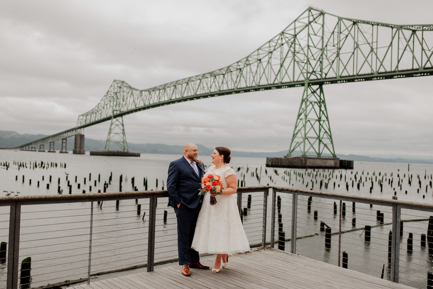 A couple in wedding attire poses on a pier with a large bridge in the background under a cloudy sky, capturing memorable photos on their special day at The Loft at the Red Building.