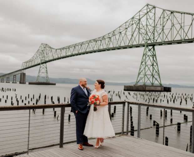 A couple poses for wedding photos on a deck by the water, with the woman holding a bouquet. A large bridge is visible in the background, marking their special day at The Loft at the Red Building.