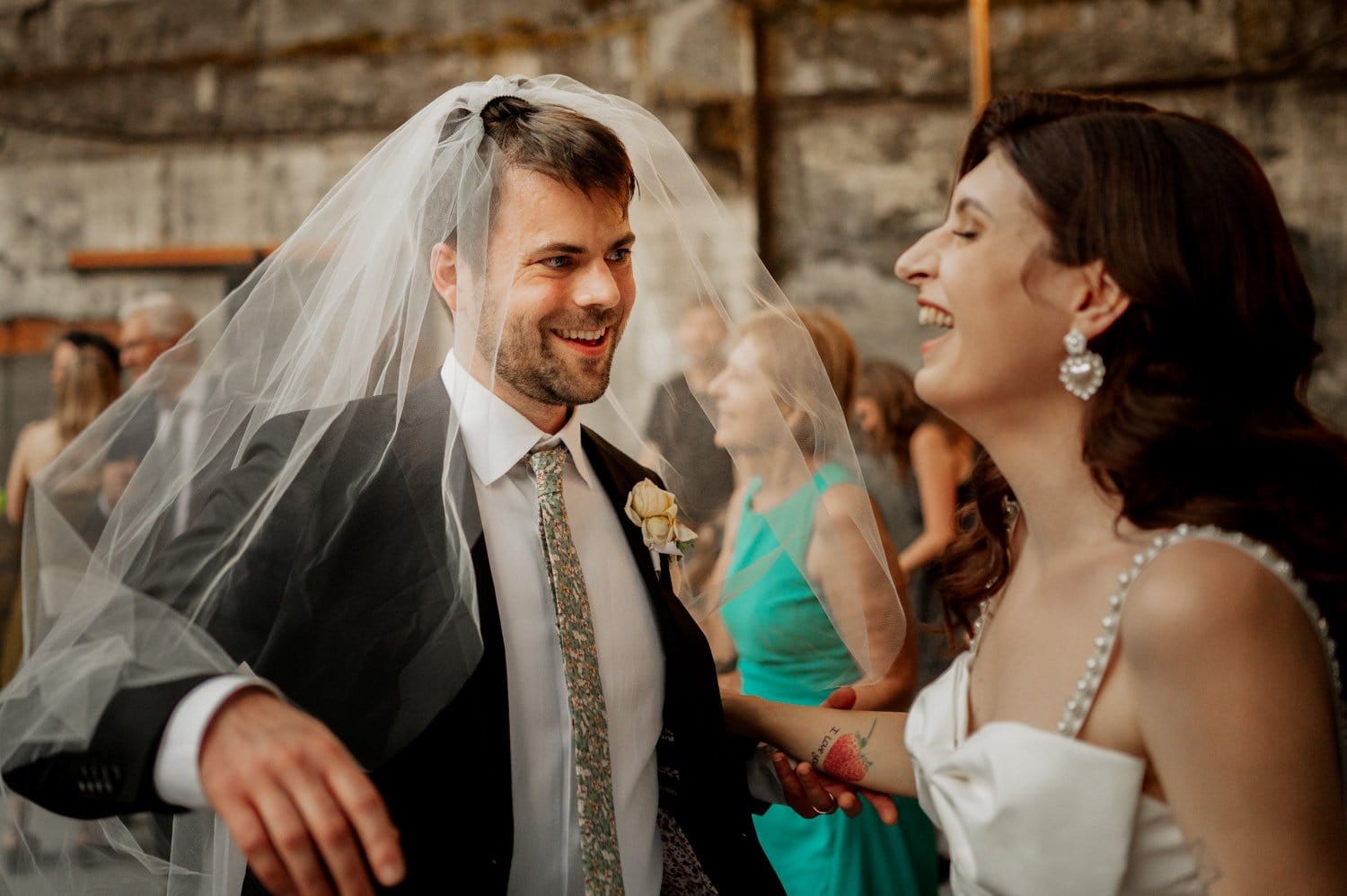 At The Ruins in Hood River, a groom playfully dons a bridal veil, beaming at his bride, who laughs joyously. Theyre elegantly dressed in wedding attire, the groom in a suit and the bride in a white gown. Blurred guests celebrate merrily in the background.