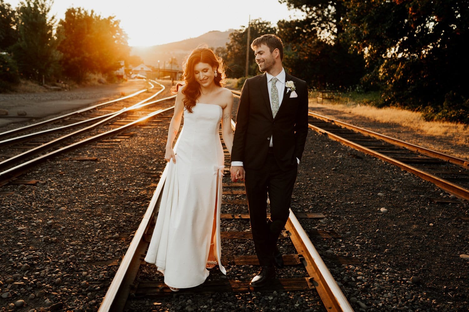 A bride in a white dress and groom in a dark suit and tie walk hand in hand on train tracks at sunset. The sun casts a warm glow, highlighting the rural scenery with trees and hills in the background.