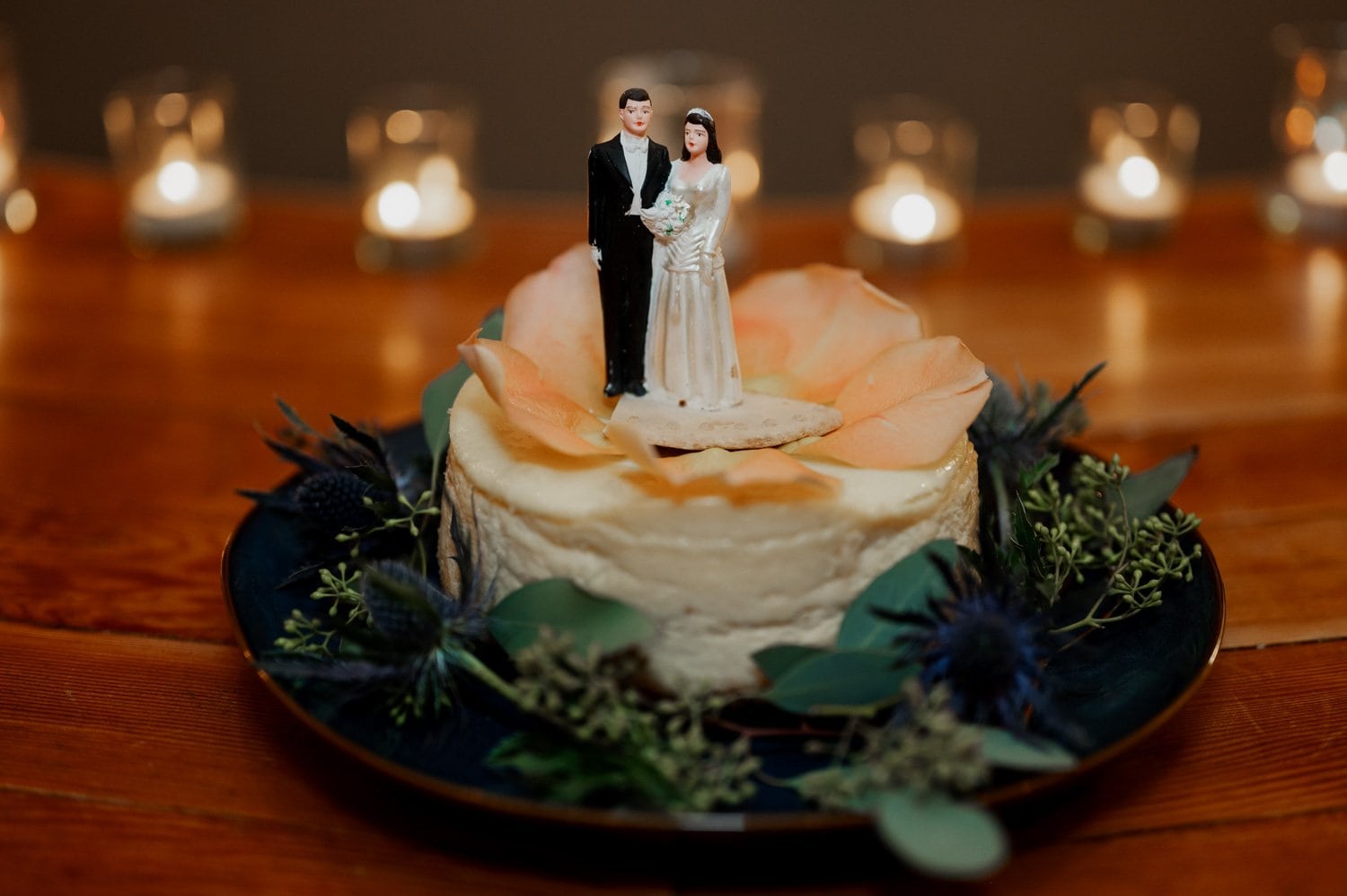 A wedding cake with a bride and groom topper on a wooden table, surrounded by candles and greenery at Opal 28.