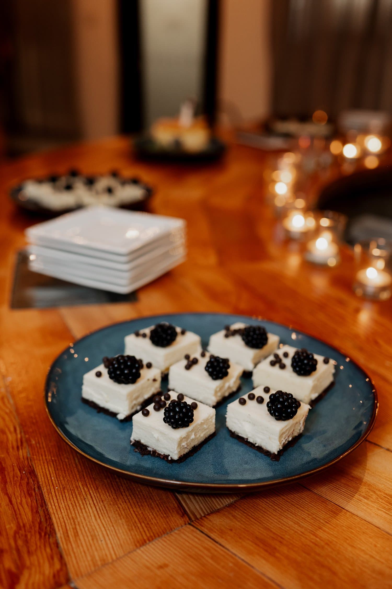Square desserts topped with cream and blackberries are neatly arranged on a blue plate. The plate is placed on a wooden table, which features small lit candles, creating an intimate atmosphere suitable for a cozy wedding at Opal 28.