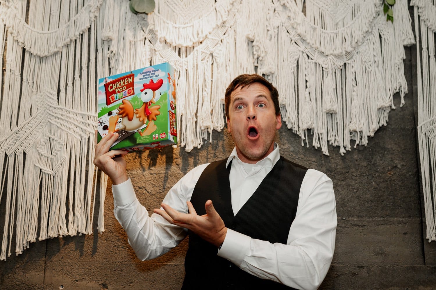 A man happily holds a colorful board game in front of a textured wall with white fringe decorations, capturing the lively atmosphere of a wedding at Opal 28.