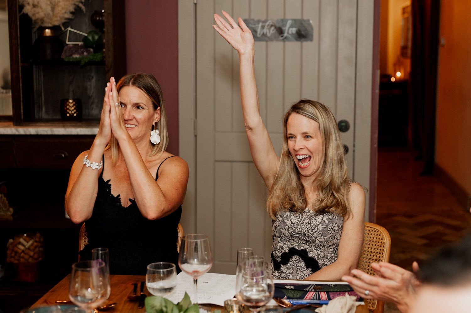 Two women sitting at a table during a wedding at Opal 28, one clapping joyfully and the other raising her hand, both with wide smiles.