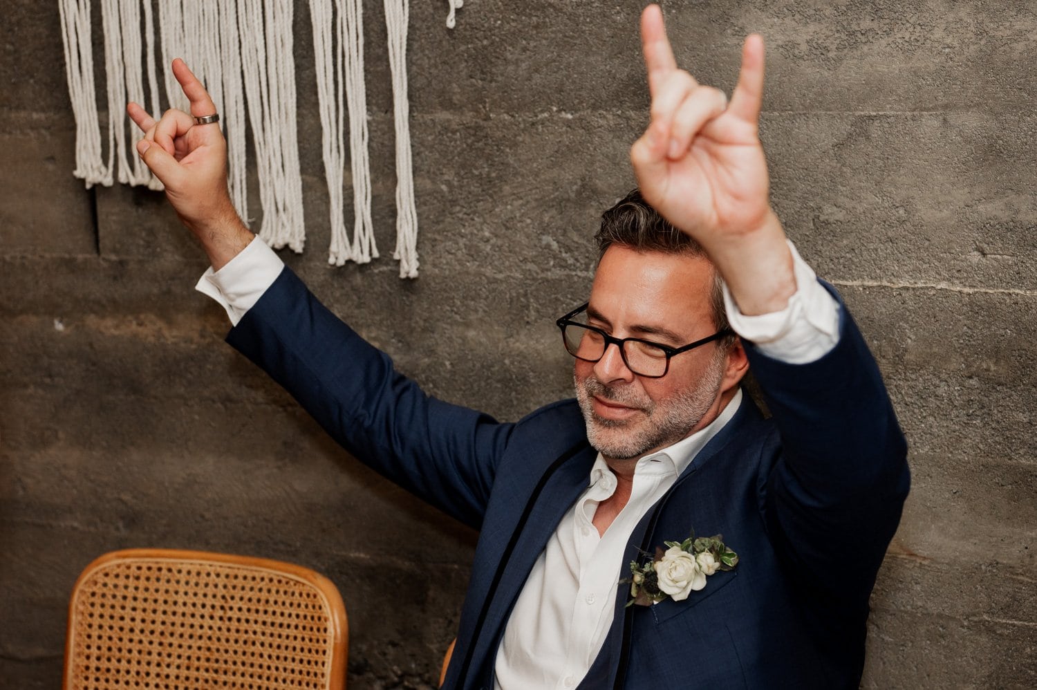 A man in a suit and glasses joyfully raises both hands, making rock signs, as he celebrates at Opal 28. A textured wall is visible behind him.