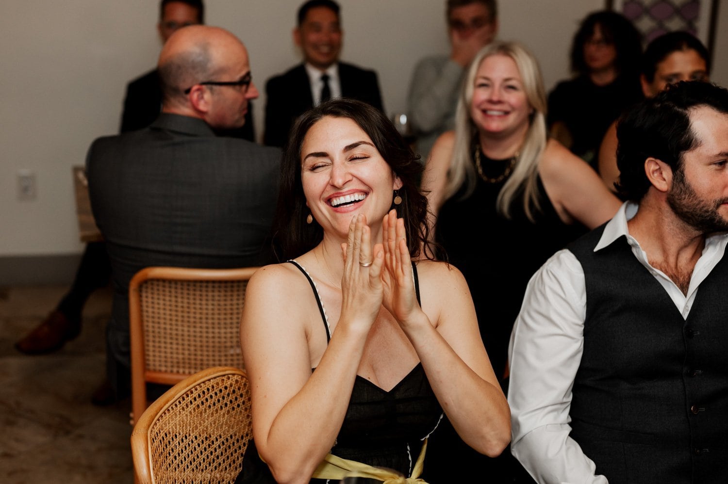 A woman in a black dress is laughing and clapping amid a wedding celebration at Opal 28, surrounded by seated guests. Her joyful expression captures the vibrant ambiance of love and togetherness.