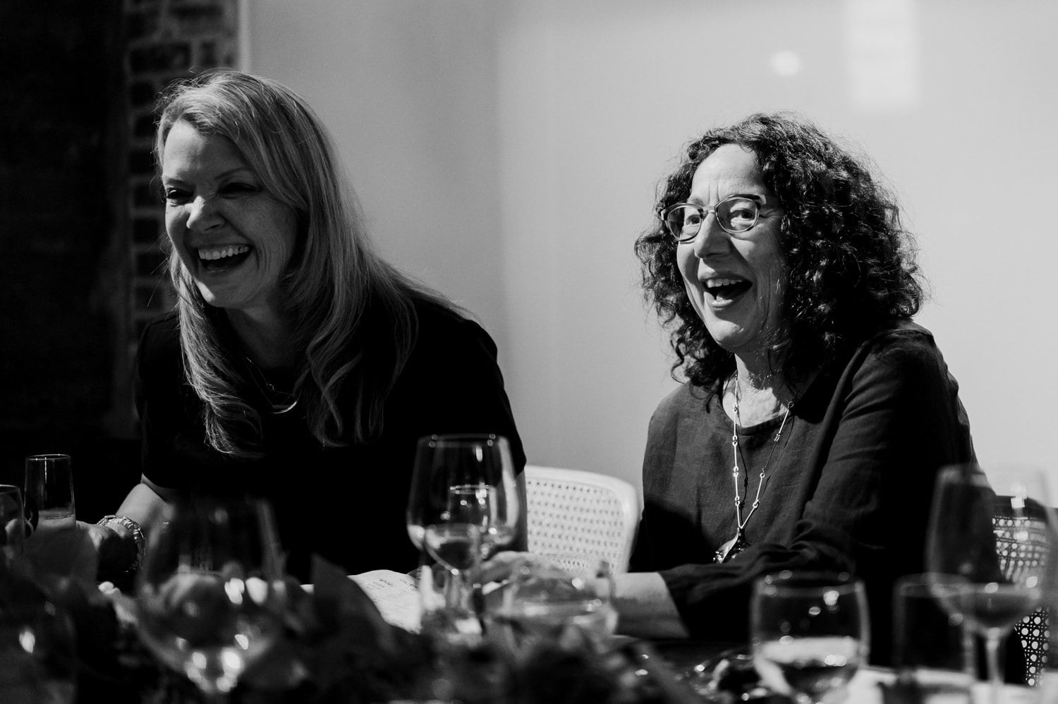 Two women sit at a table, laughing with glasses of wine in hand and plates in front of them, captured in a black and white photo.