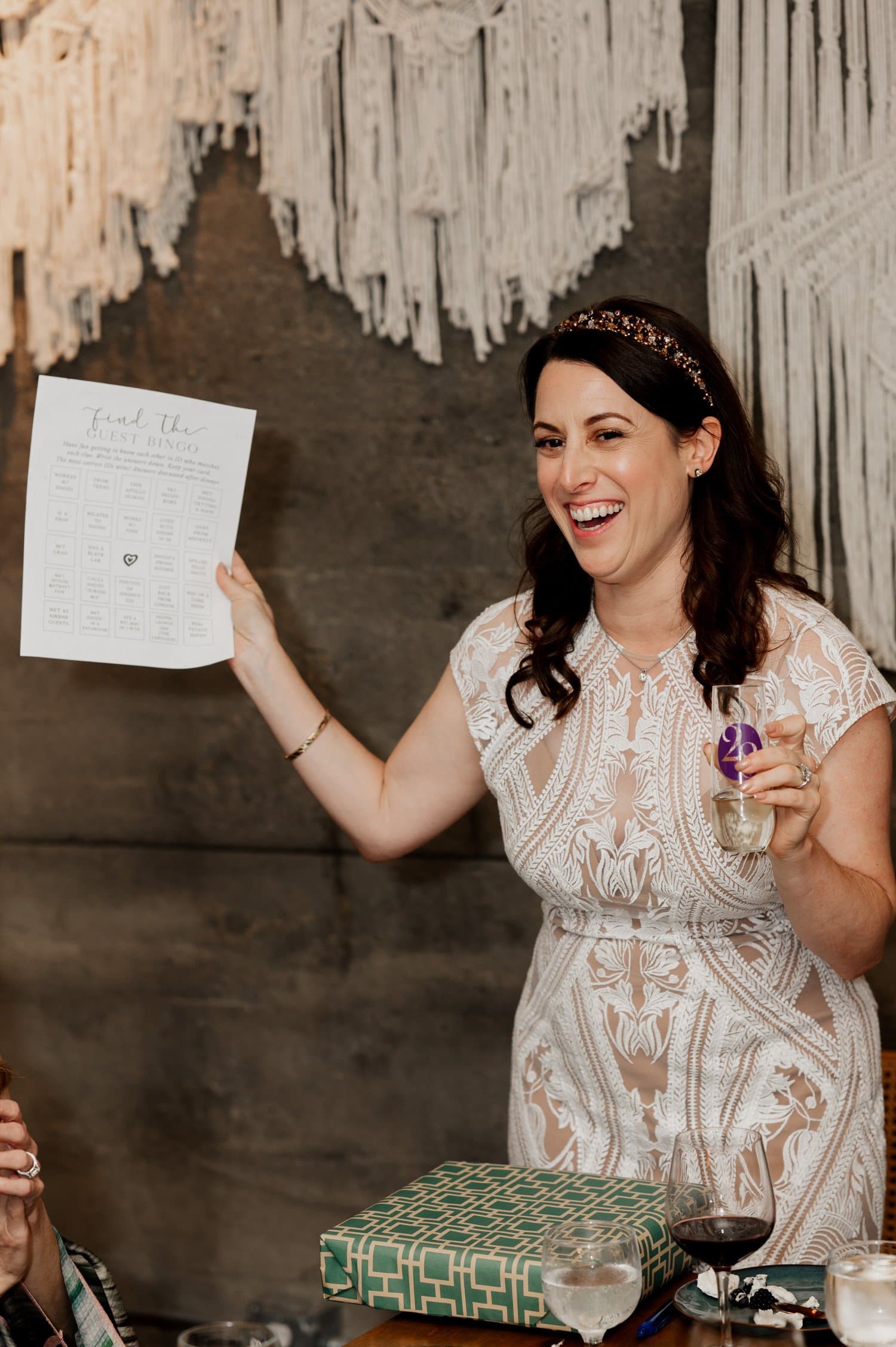 A woman smiles while holding a bingo card and a drink. A wrapped gift is on the table in front of her. The background features macrame decorations at Opal 28 during a wedding event.