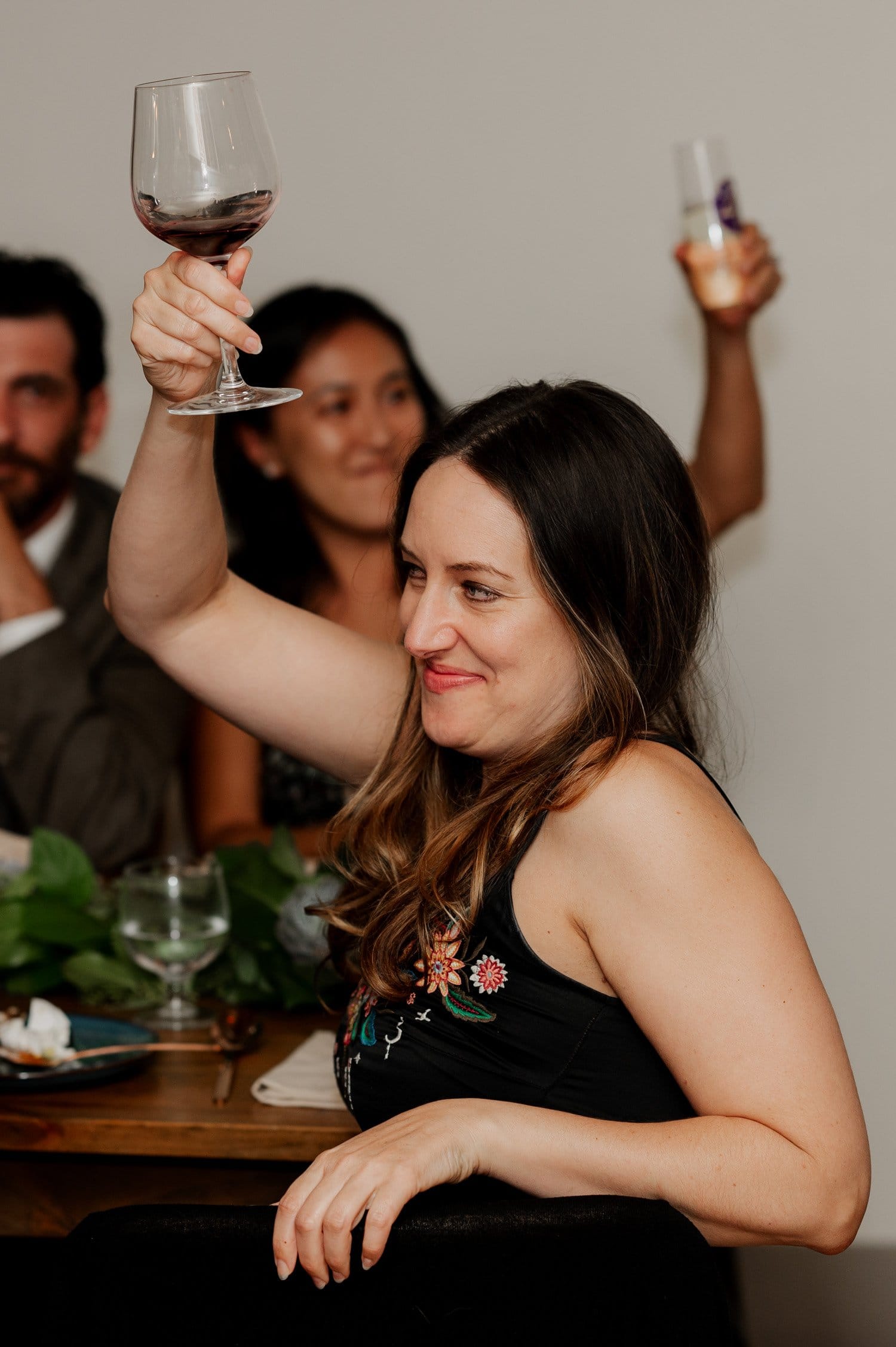 A woman in a formal dress raises her wine glass in a toast at a wedding, smiling brightly. Two guests stand in the background, holding drinks and participating in the celebration.