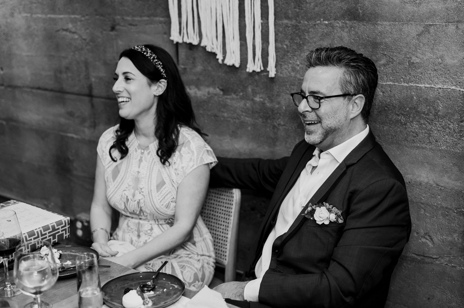 A smiling couple sits together at a table, with the man in a suit featuring a boutonniere, in the warmly lit room of their wedding at Opal 28.