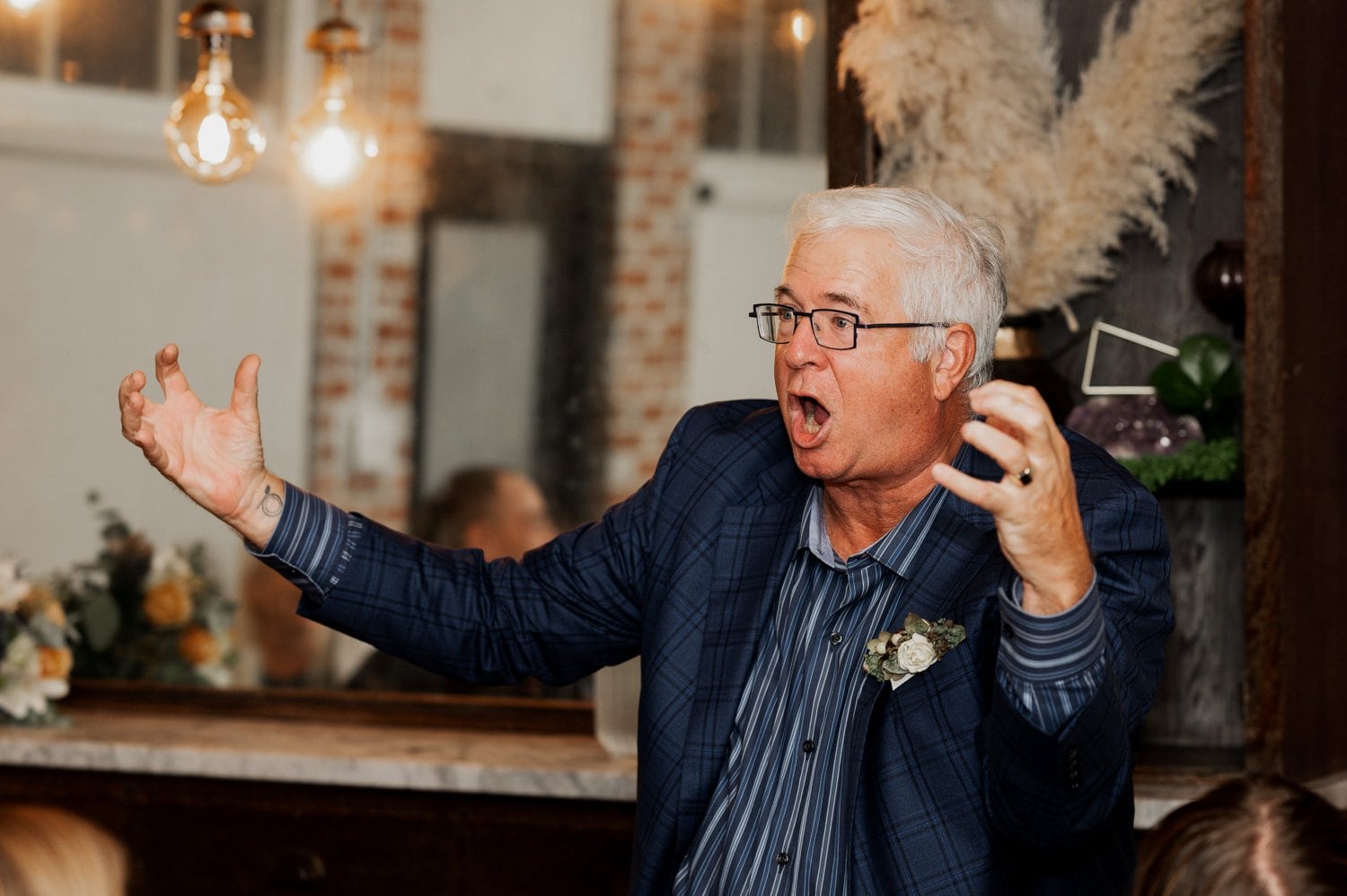 A man with glasses passionately speaks and gestures with open hands in a dimly lit room, wearing a blue plaid suit jacket.
