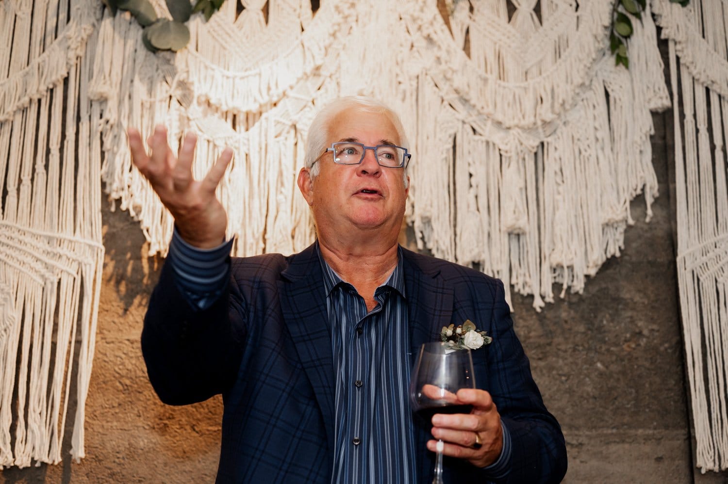 A man in a suit gracefully gestures with one hand while enjoying a glass of wine, standing in front of an ornate macramé backdrop at a wedding at Opal 28.