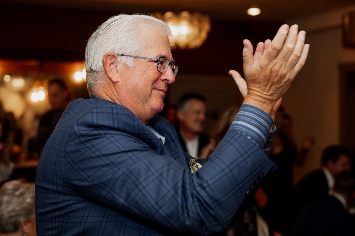 An elderly man in a blue plaid suit claps enthusiastically, surrounded by other guests celebrating in a warmly lit venue at Opal 28 during a wedding.