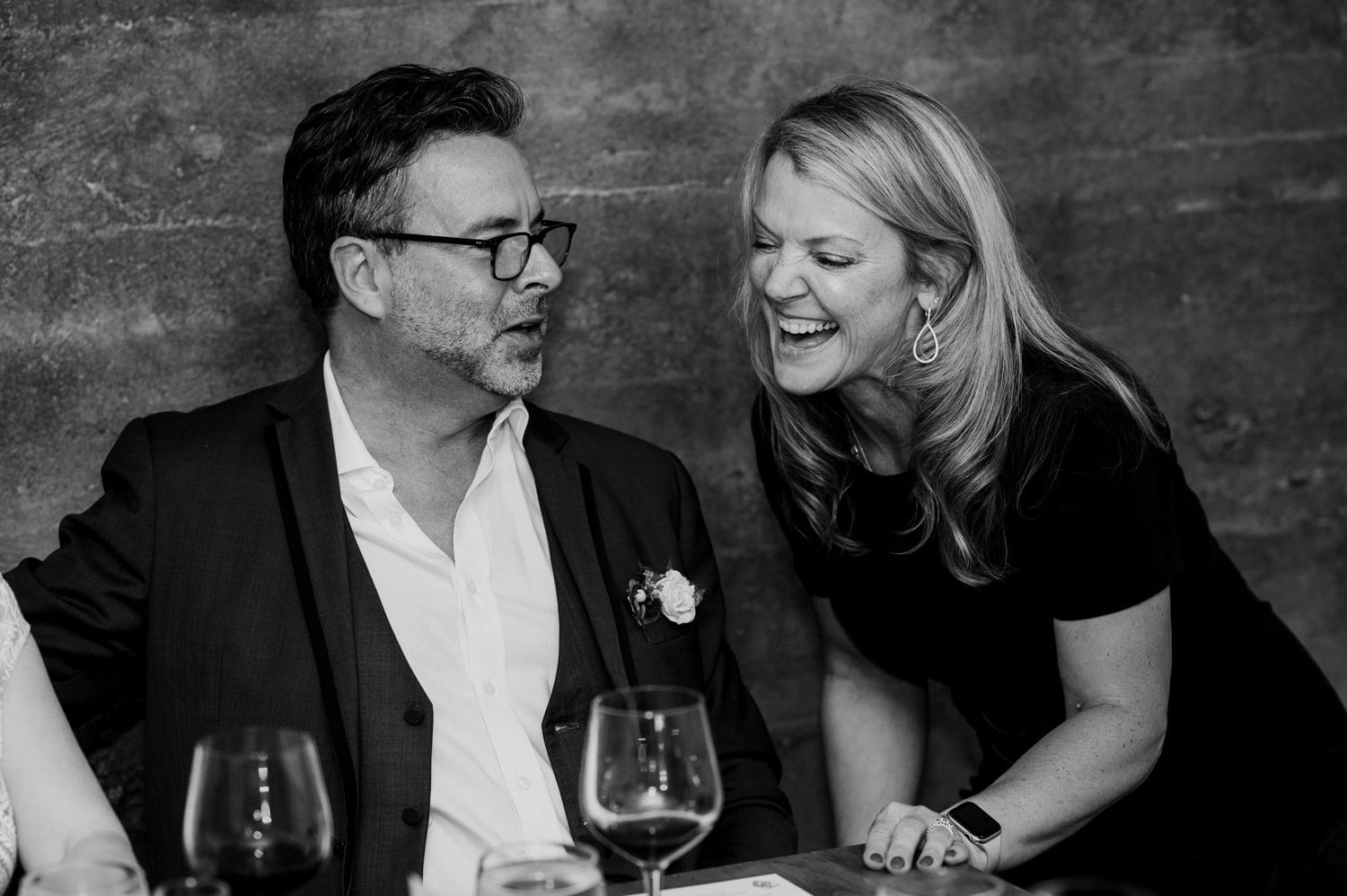 A man in a suit and a woman in a black dress are laughing together at a table with wine glasses, enjoying a wedding celebration at Opal 28.