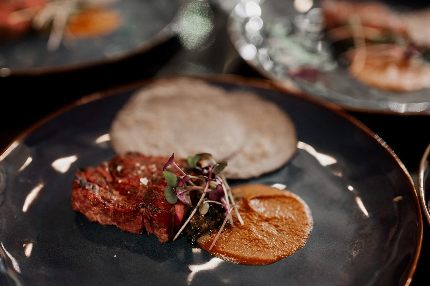 A gourmet steak dish with sauce and microgreens is presented on a dark plate, surrounded by other elegant meals, suitable for a wedding celebration at Opal 28.