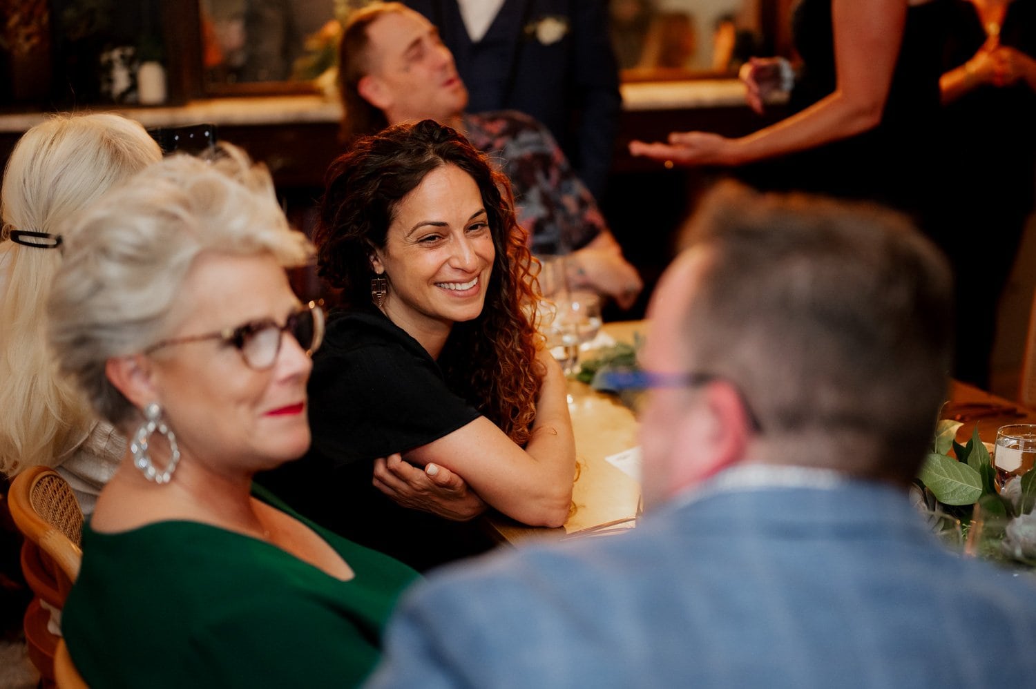 A group of people sits around a table at a wedding at Opal 28, engaging in conversation and smiling. Some guests are holding drinks, while the table is decorated with flowers and candles. Everyone is dressed in formal attire, creating a festive atmosphere.