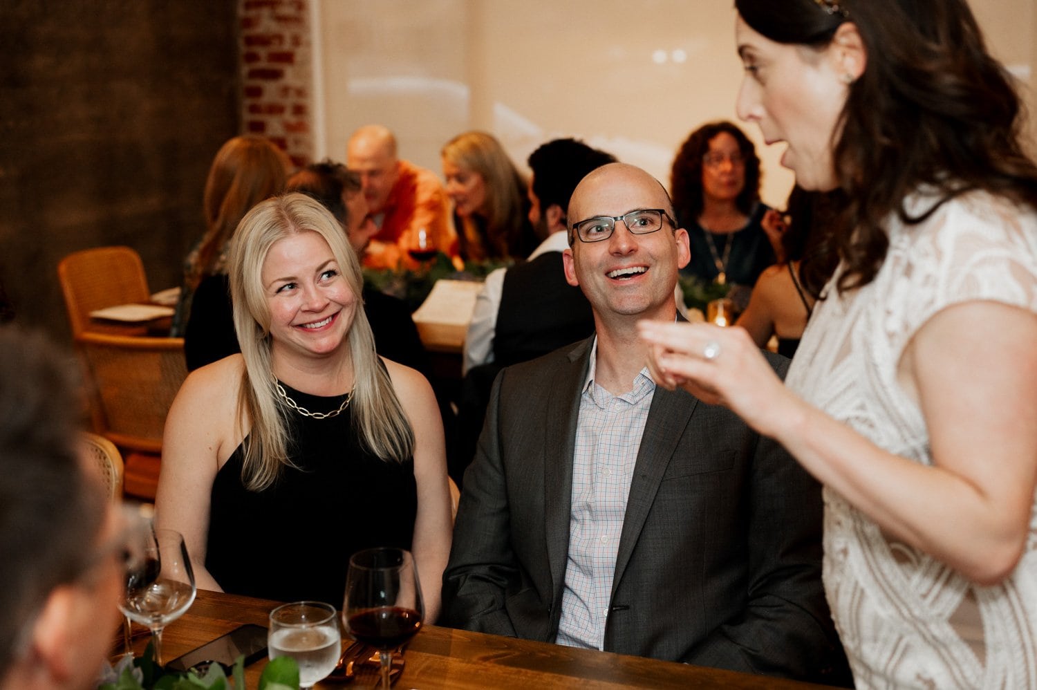 Guests engage in animated conversations around a table at Opal 28, while others stand nearby with drinks in hand, all contributing to a joyful wedding atmosphere.