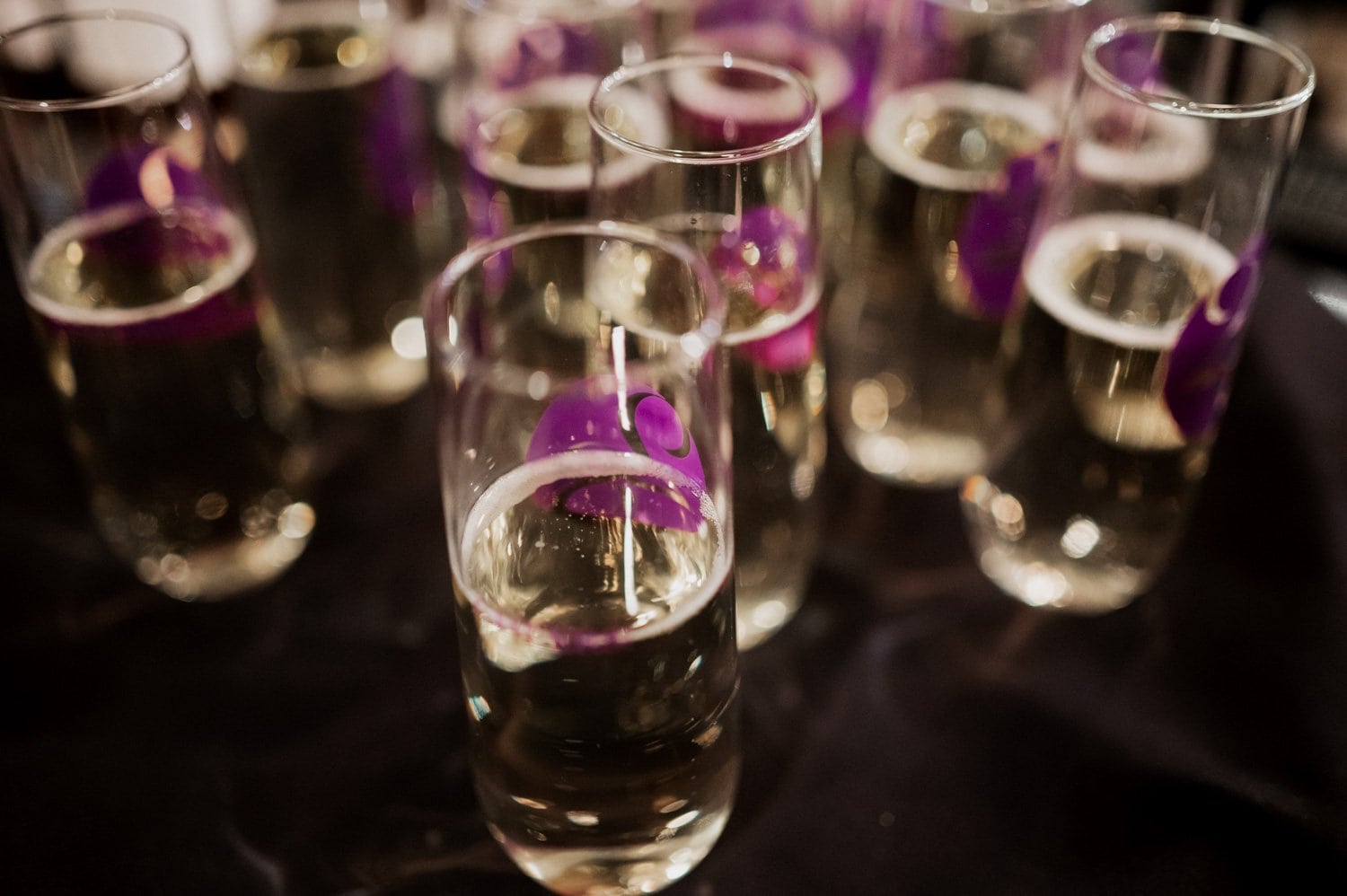 Glasses of champagne with small purple flower garnishes on a dark table at a wedding reception.