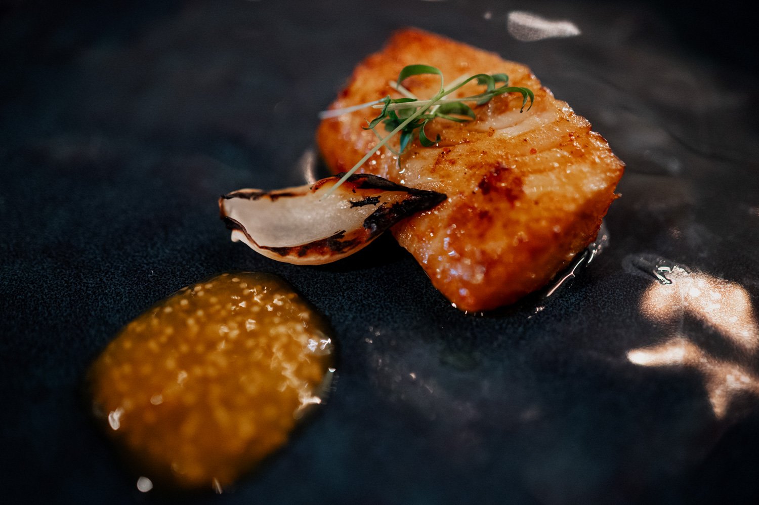 Grilled fish topped with a charred onion slice and microgreens, accompanied by mustard sauce on a dark plate. Ideal for an elegant wedding at Opal 28.