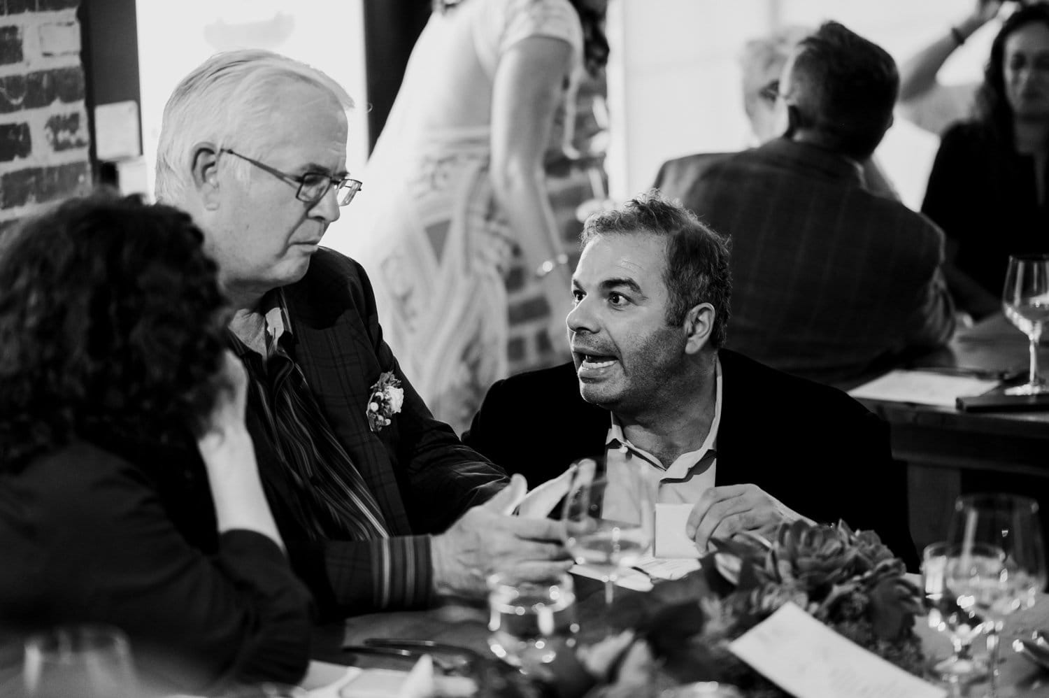 Two men talking at a table in a lively indoor setting, with people milling around them. The scene is captured in black and white and suggests a wedding gathering at Opal 28.