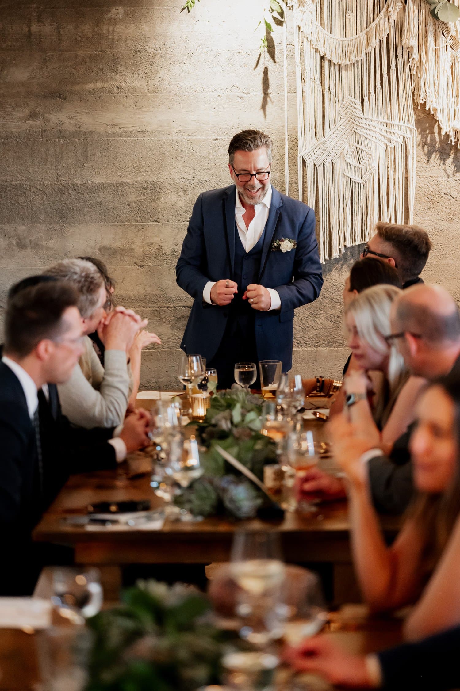 A man in a blue suit speaks at a wedding dinner at Opal 28, with guests seated around a table decorated with a macrame hanging.
