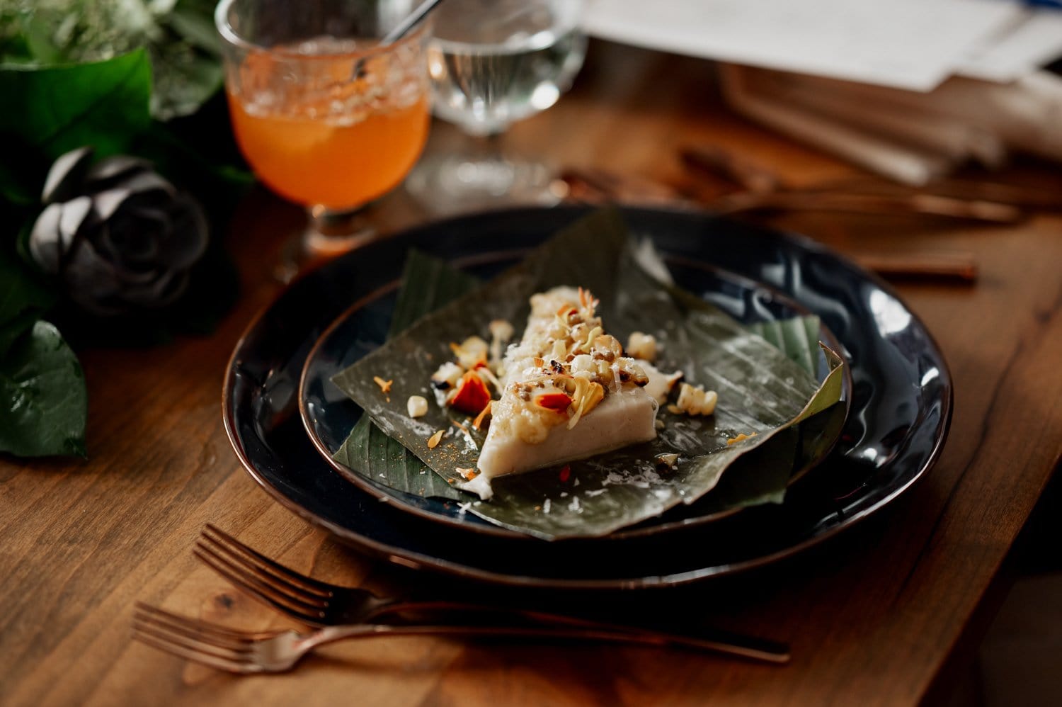 A dessert wrapped in a leaf, garnished with nuts and seeds, is placed on a black plate beside a glass of orange juice on an elegant table setting.