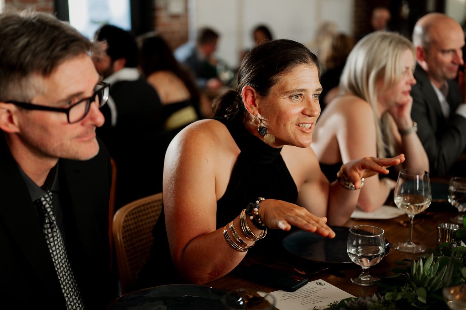 At a lively dining table during a wedding at Opal 28, a woman energetically gestures while talking with several guests.