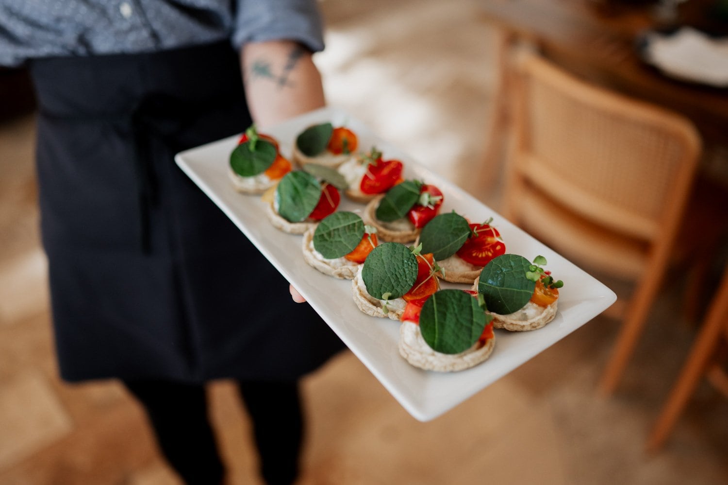 A person holds a tray with an elegant white plate featuring crackers topped with ripe tomatoes and green leaves, set against the backdrop of a charming wedding at Opal 28.
