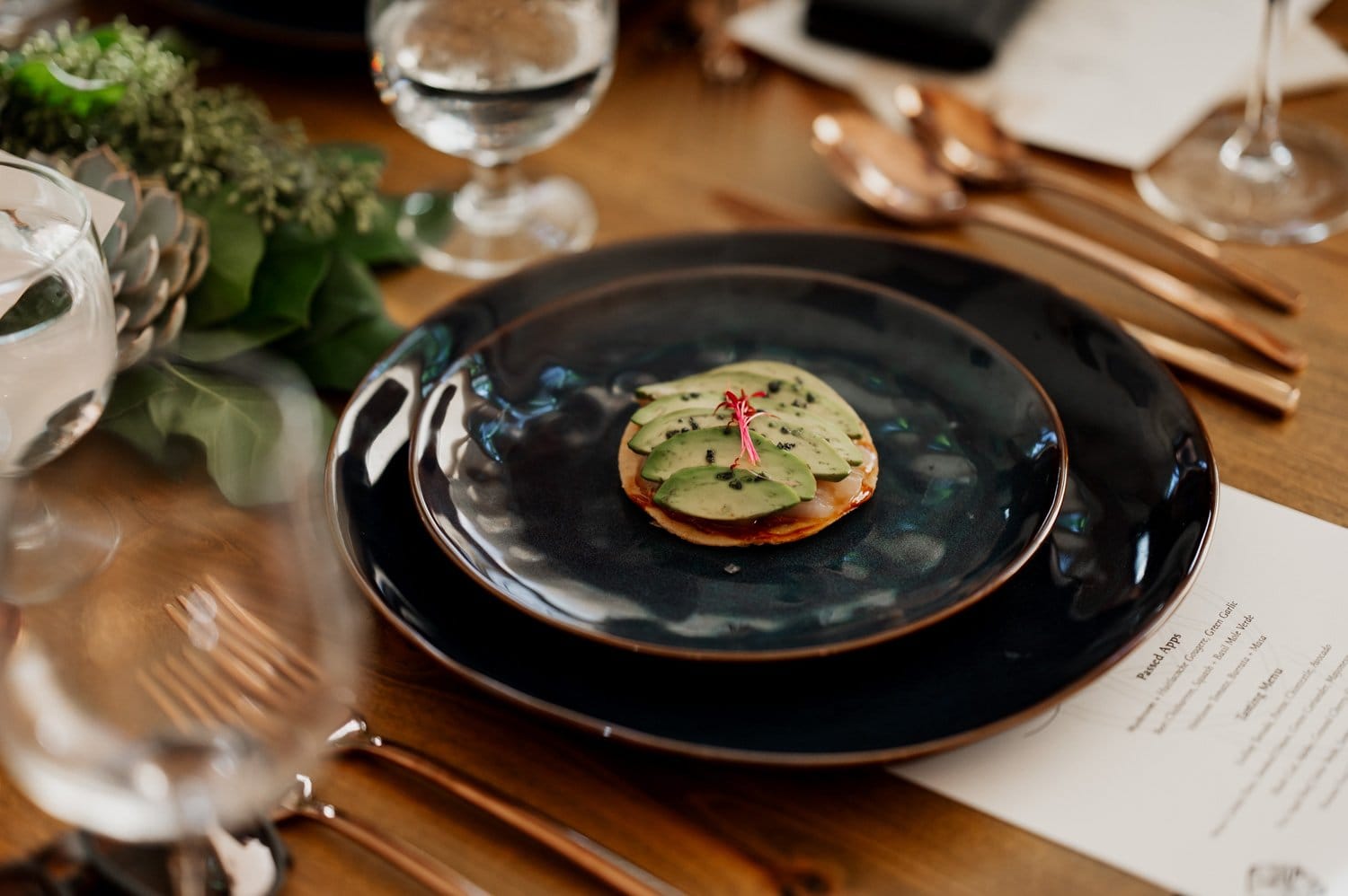 An elegantly arranged table setting featuring a small gourmet appetizer on a dark plate, with neatly placed cutlery and sparkling water glasses surrounding it, prepared for a wedding at Opal 28.